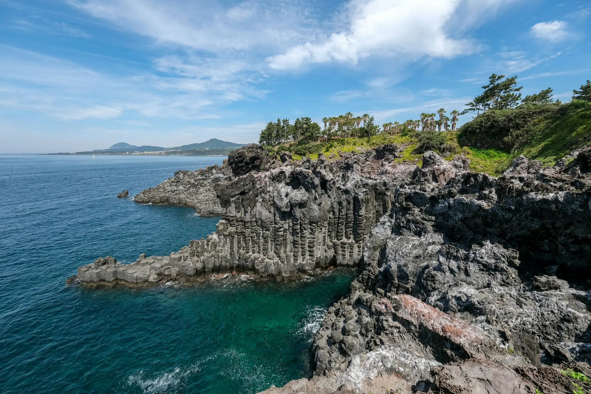 Columnar joint cliffs and turquoise sea at Jusangjeolli Cliff in Jeju Island, South Korea, under a clear blue sky – a top natural attraction for family and Muslim-friendly travel itineraries.