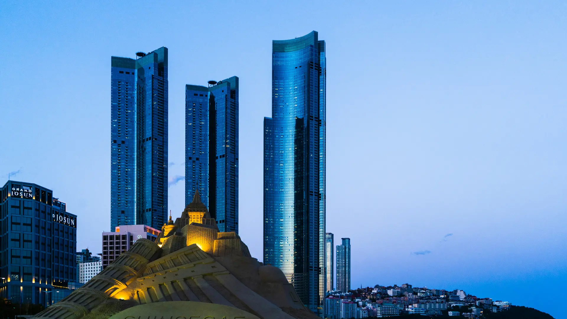 A wide-angle landscape shot of Haeundae Beach in Busan at dusk. In the foreground, a large, intricately carved sandcastle sculpture is illuminated by warm spotlights, showing detailed architectural features like towers and windows.  Behind the sculpture, the skyline is dominated by three glowing, blue-tinted glass skyscrapers (Haeundae LCT The Sharp) that stretch toward a deep blue twilight sky. To the left, the "Grand Josun" hotel building is visible, and to the right, the city lights of a hillside neighborhood begin to twinkle. The overall mood is calm and sophisticated, contrasting the organic texture of the sand with the sleek, modern urban architecture.