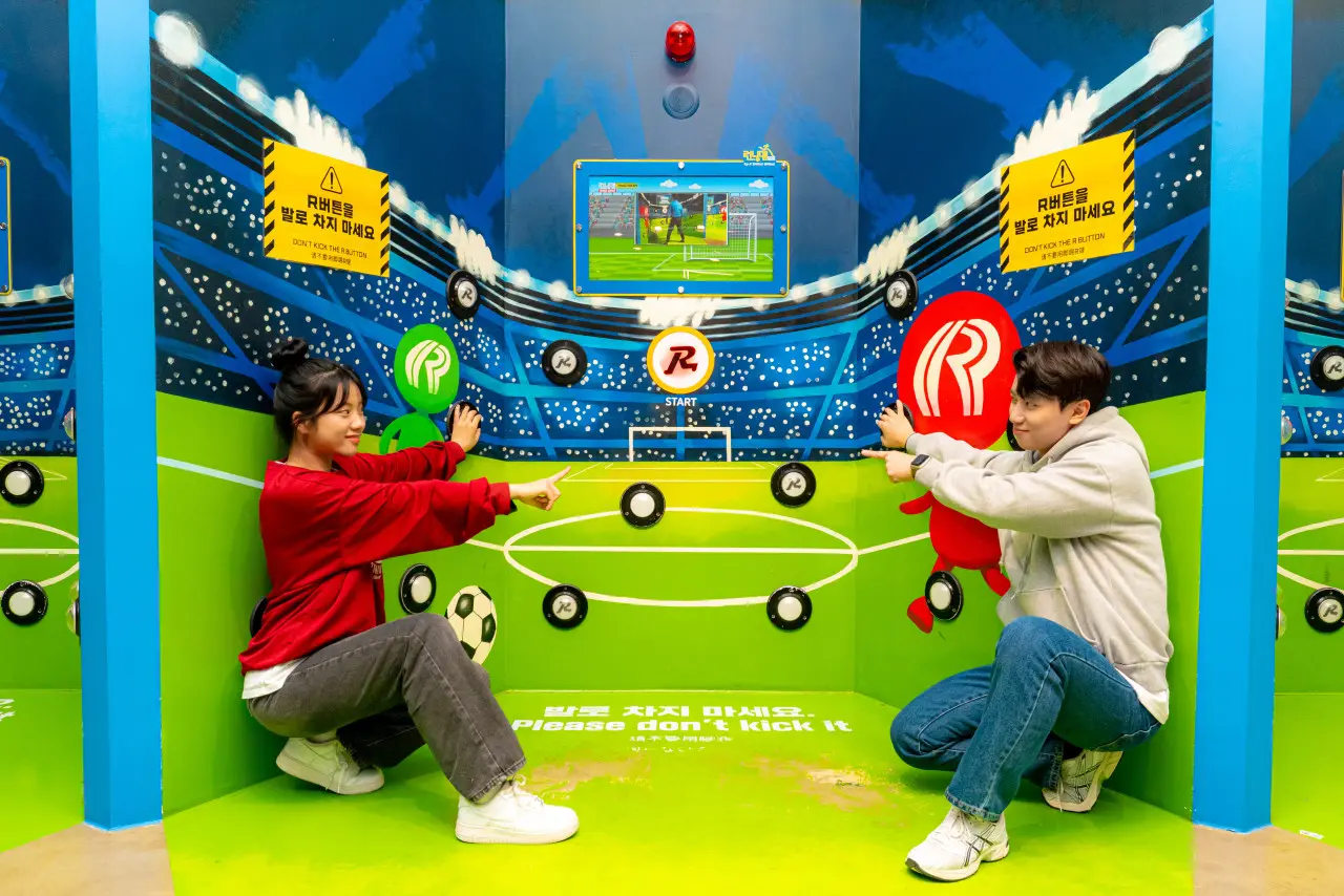 A vibrant, eye-level shot of an interactive game zone at Running Man Busan. Two people, a woman in a red sweatshirt and a man in a grey hoodie, are crouched on a bright green floor designed to look like a soccer field. They are both pointing playfully toward a central "START" button featuring the iconic Running Man "R" logo.  The walls are decorated with a colorful stadium-themed mural, complete with illustrated crowds and soccer goals. Several black circular touch-pads are scattered across the walls as part of the game. Two yellow warning signs in Korean and English advise players, "Please don't kick it." In the center, a digital screen displays a soccer-themed mini-game, and stylized red and green "R" character figures flank the players, adding to the energetic and immersive atmosphere of the attraction.