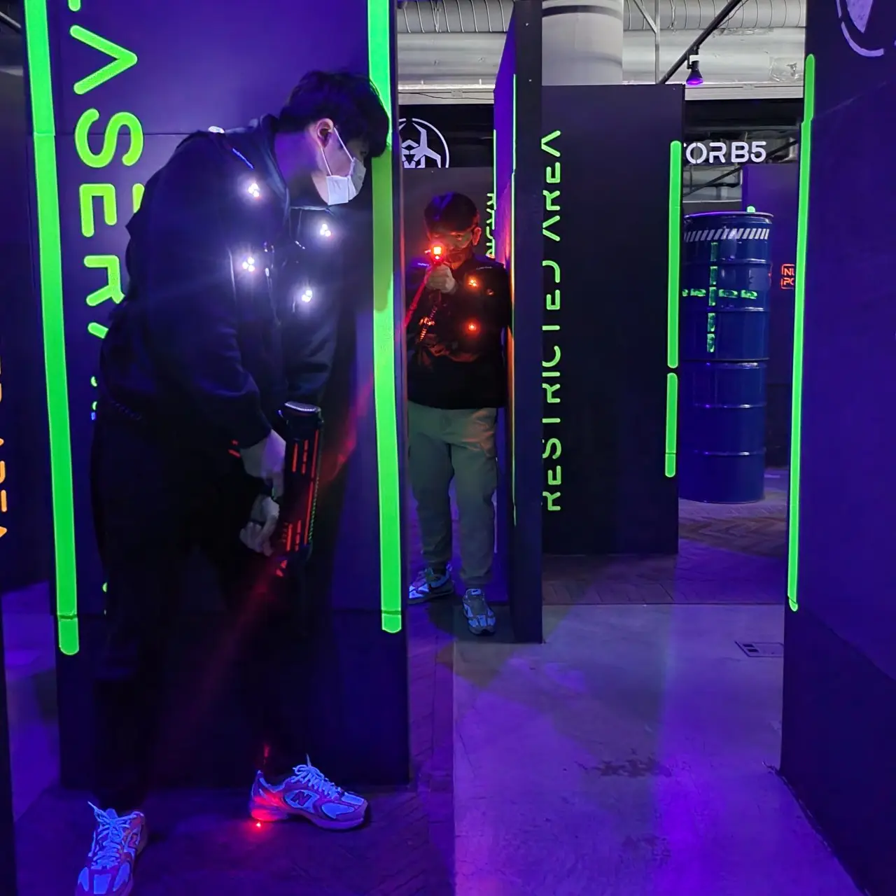 A medium-shot of two people engaged in a laser tag game at Laser Arena Busan Samjeong. The setting is a dark, indoor arena illuminated by vibrant neon green lights on black partition walls labeled with text like "RESTRICTED AREA."  In the foreground, a person wearing a black hoodie, a face mask, and glowing sensors on their vest stands in a defensive posture, holding a laser gun. In the background, another player is partially visible behind a partition, aiming a laser gun that emits a bright red beam toward the foreground. The floor has a purple hue from the ambient neon lighting, creating an intense, high-energy atmosphere for an indoor action activity.
