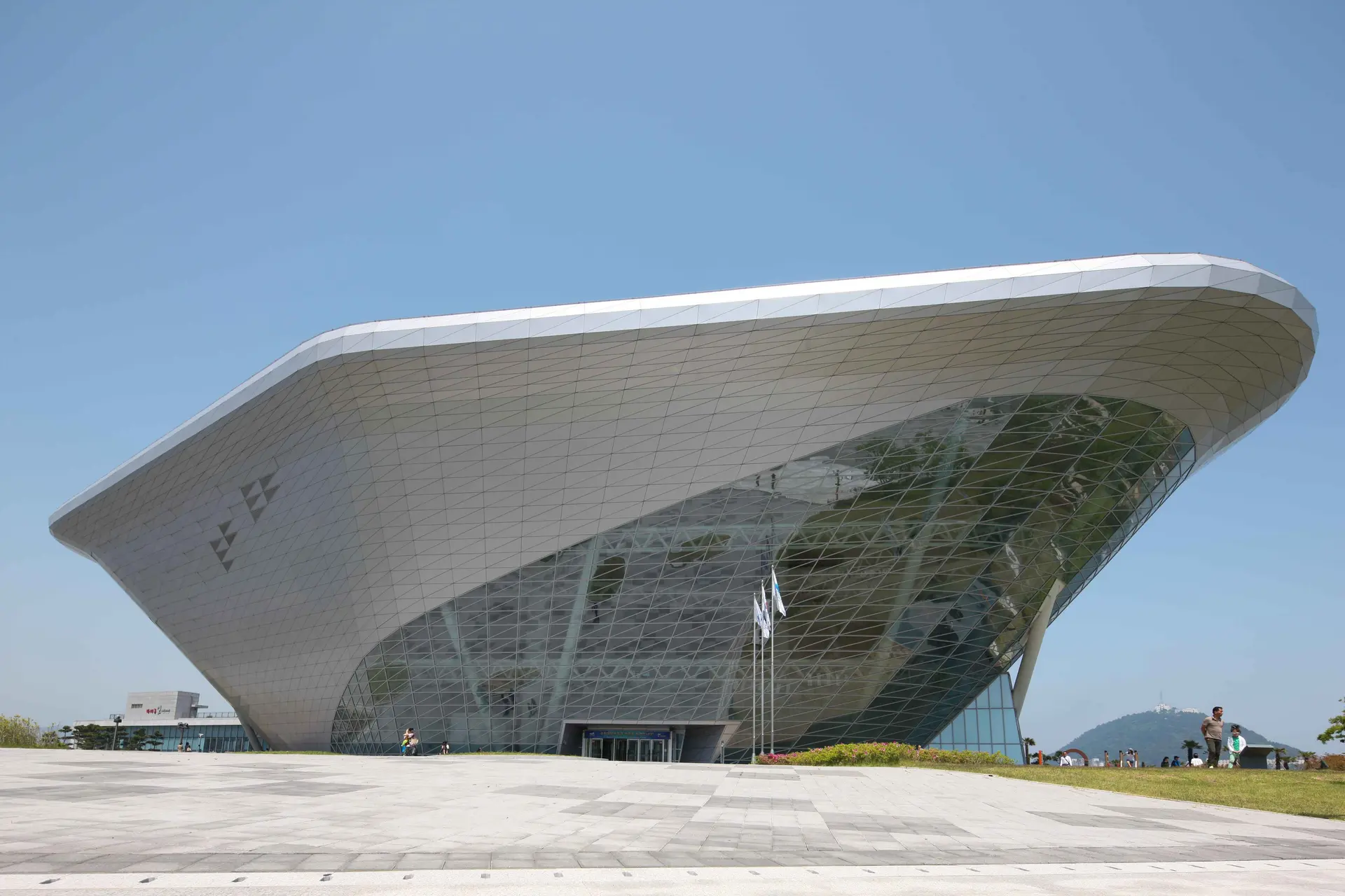  A wide-angle, low-angle shot of the Korea National Maritime Museum in Busan. The building features a striking, futuristic architectural design with a large, silver metallic canopy that flares upward and outward, resembling a stylized ship's hull or an inverted pyramid. The facade is covered in a grid of triangular glass panels that reflect the surrounding environment.  In the foreground, a vast, light-gray paved plaza stretches toward the entrance. A few small figures of visitors are scattered near the building and on the grassy area to the right, highlighting the massive scale of the structure. The sky is a clear, pale blue, and a distant, hazy mountain is visible on the horizon to the right, completing the serene coastal setting.