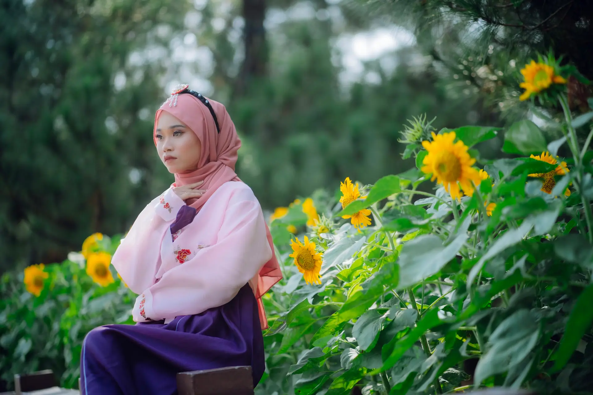 A profile shot of a woman wearing a traditional Korean Hanbok and a pink hijab, seated on a wooden bench. She is dressed in a light pink jeogori (top) with delicate red floral embroidery and a deep purple chima (skirt). She wears a decorative traditional headpiece over her hijab.  She is positioned on the left side of the frame, looking off-camera with a serene expression. To her right, a cluster of vibrant yellow sunflowers is in full bloom, with lush green leaves catching the sunlight. The background is a soft, blurred bokeh of green trees and foliage, creating a peaceful and graceful outdoor setting.
