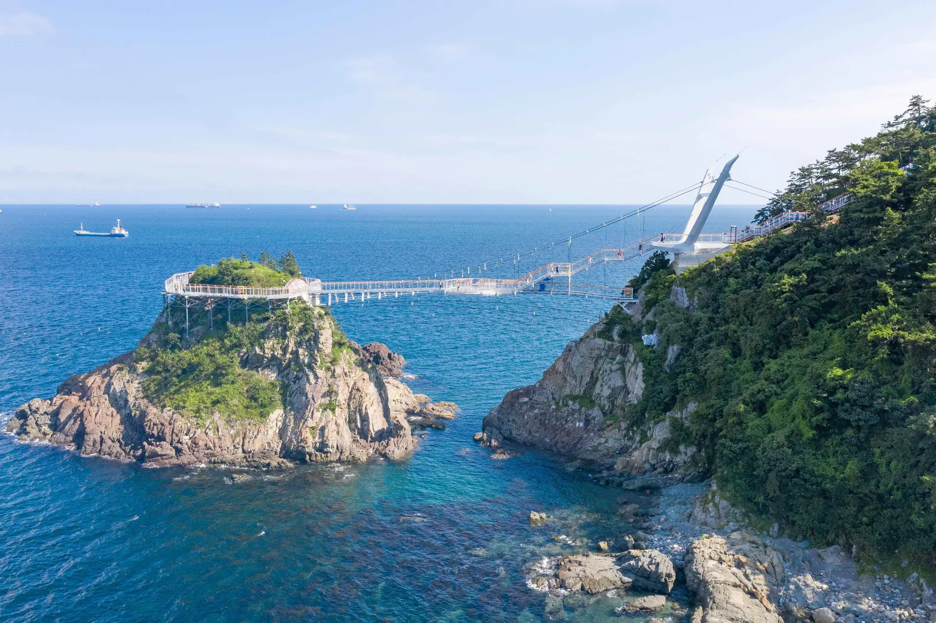 A wide-angle, high-angle shot of the Songdo Yonggung Suspension Bridge in Busan. The white metal bridge stretches across a turquoise sea, connecting the mainland cliff on the right to a small, rocky uninhabited island on the left.  The island features a circular walking path at its peak, surrounded by sparse greenery. The mainland side is a steep, lush green hillside with a white observation tower at the bridge's starting point. In the background, the deep blue ocean extends to the horizon, where several large cargo ships are visible under a clear, pale blue sky. The scene highlights the dramatic coastal geography and the engineering of the pedestrian bridge.