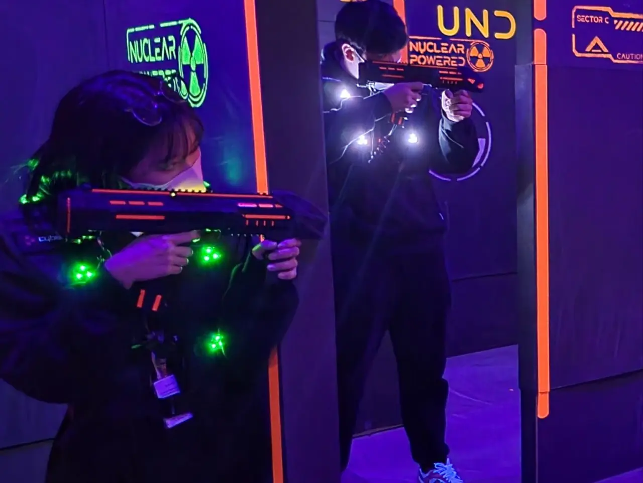 A medium-shot of two people engaged in a laser tag game at Laser Arena Busan. The setting is a dark, indoor arena illuminated by vibrant neon orange and green lights on black partition walls featuring tactical signage like "NUCLEAR POWERED" and "SECTOR C."  In the foreground, a person wearing a black jacket and a face mask aims a laser gun that glows with orange lights. Their tactical vest is lit up with bright green sensor lights. Behind a central partition, another player is visible, also wearing a lit-up vest and aiming their weapon toward the camera. The floor and surroundings are bathed in a deep purple ultraviolet light, creating an intense, high-energy atmosphere for an indoor action activity.