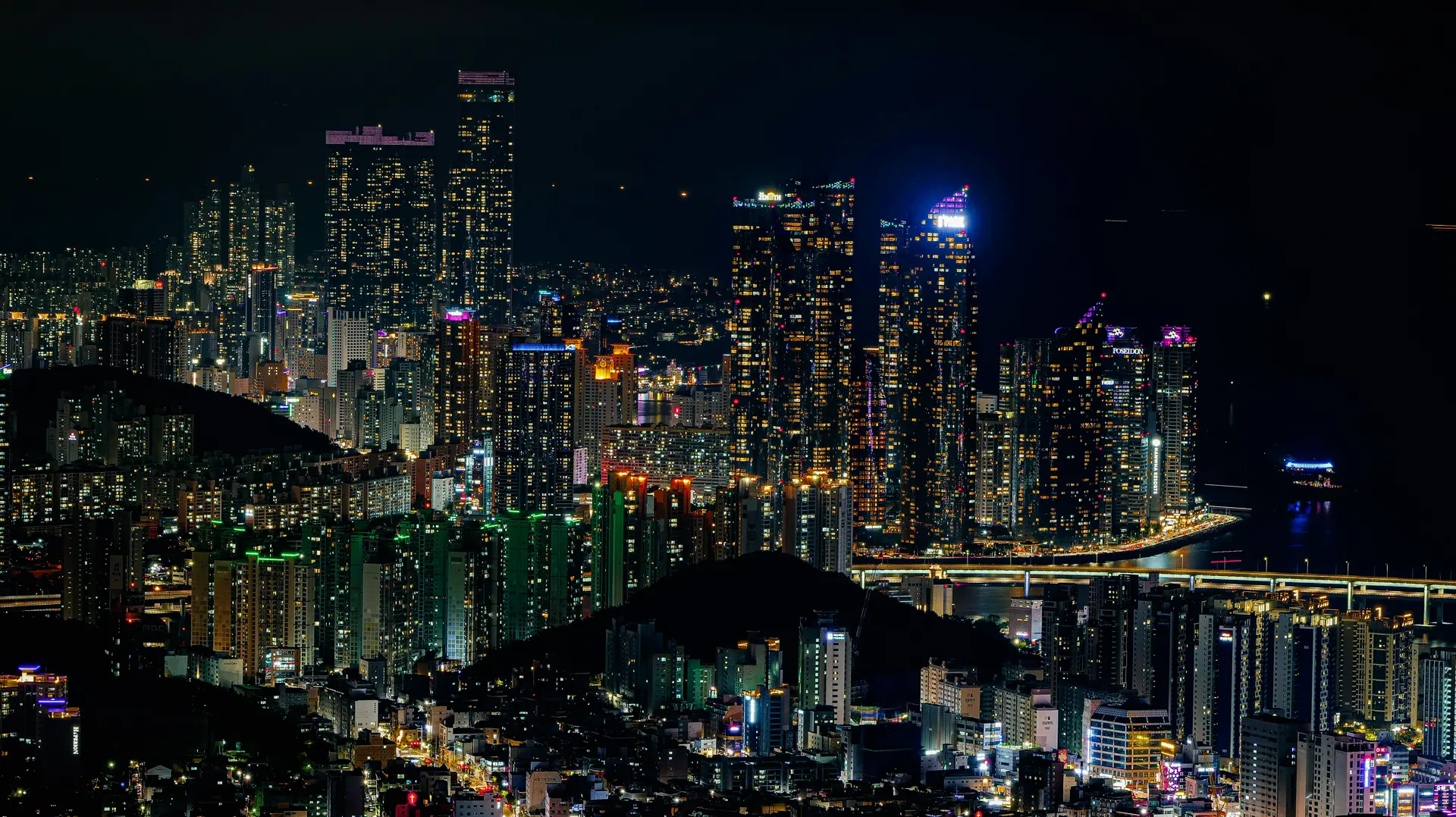  A wide-angle, low-angle landscape shot of the Marine City skyline in Haeundae, Busan, at night. The foreground features a calm, dark sea that perfectly reflects the vibrant, glowing lights of the surrounding architecture.  The skyline is dominated by several luxury high-rise skyscrapers, including the iconic curved glass towers of Haeundae I'Park and the towering Doosan We've the Zenith. The buildings are brilliantly illuminated with thousands of white and blue internal lights, creating a "festival of light" that mimics the aesthetic of major global cities like Hong Kong or Shanghai.  On the right, the Gwangan Bridge (Diamond Bridge) stretches across the water, its double-deck suspension structure glowing with bright LED lights. The sky is a deep, clear indigo, providing a sharp contrast to the shimmering gold and blue reflections dancing on the water's surface. The overall atmosphere is modern, sophisticated, and energetic, capturing Busan's status as a premier global port city.
