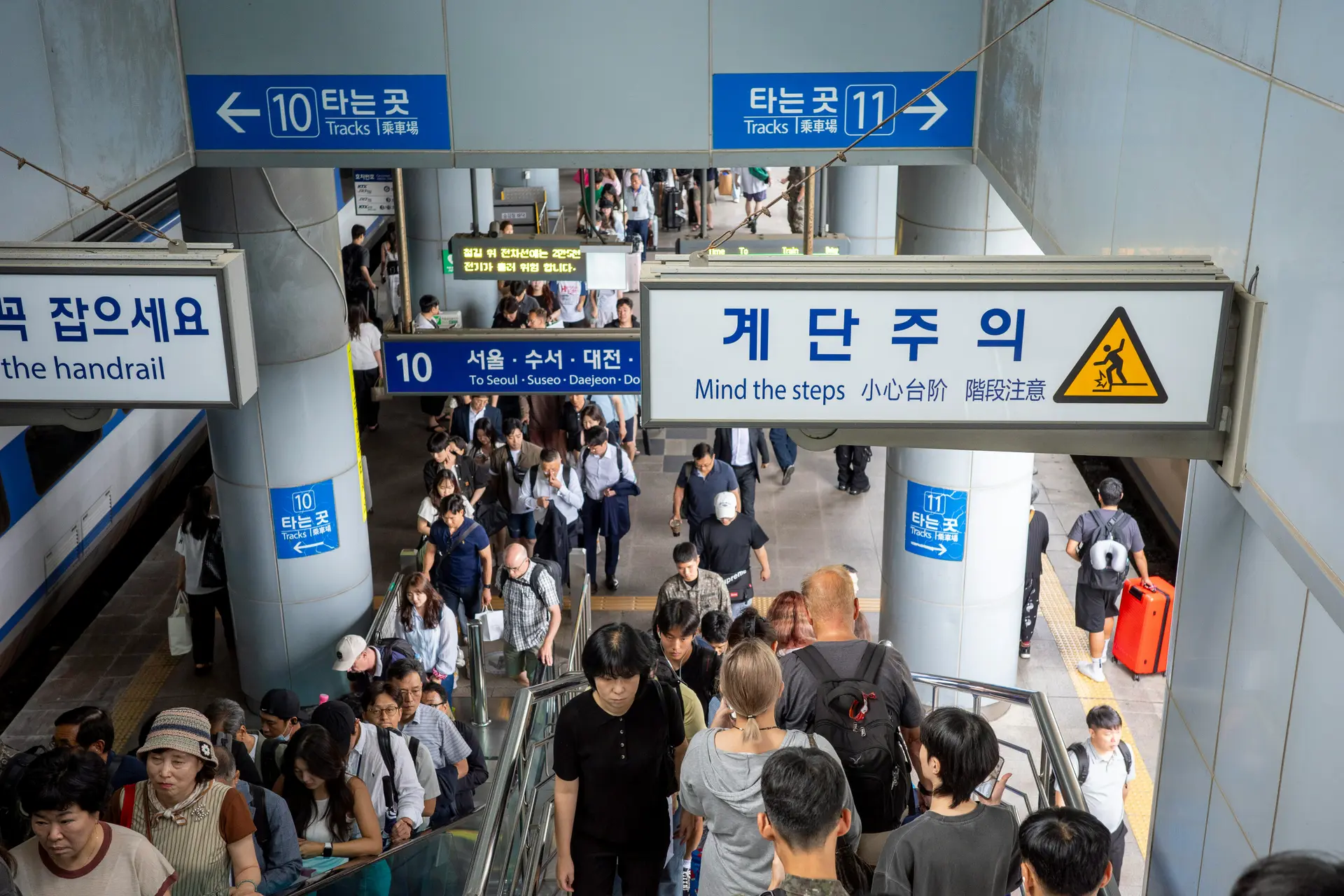 Crowded KTX station stairwell in Korea with travelers carrying luggage under blue directional signs pointing to Seoul and Daejeon, illustrating the busy Seollal holiday travel rush and high demand for intercity train tickets.