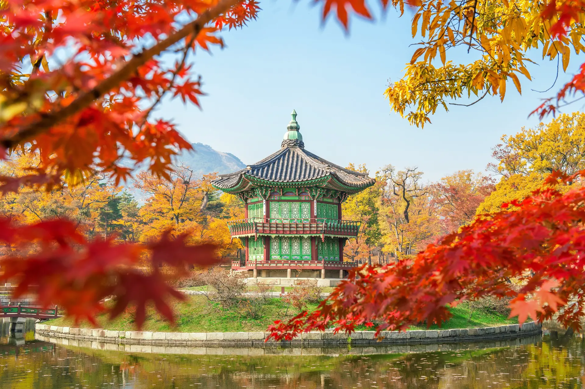 Scenic view of the hexagonal Hyangwonjeong Pavilion situated on Hyangwonji Pond within Gyeongbokgung Palace, Seoul, South Korea. The pavilion is framed by vibrant red maple leaves and yellow autumn foliage under a clear blue sky.