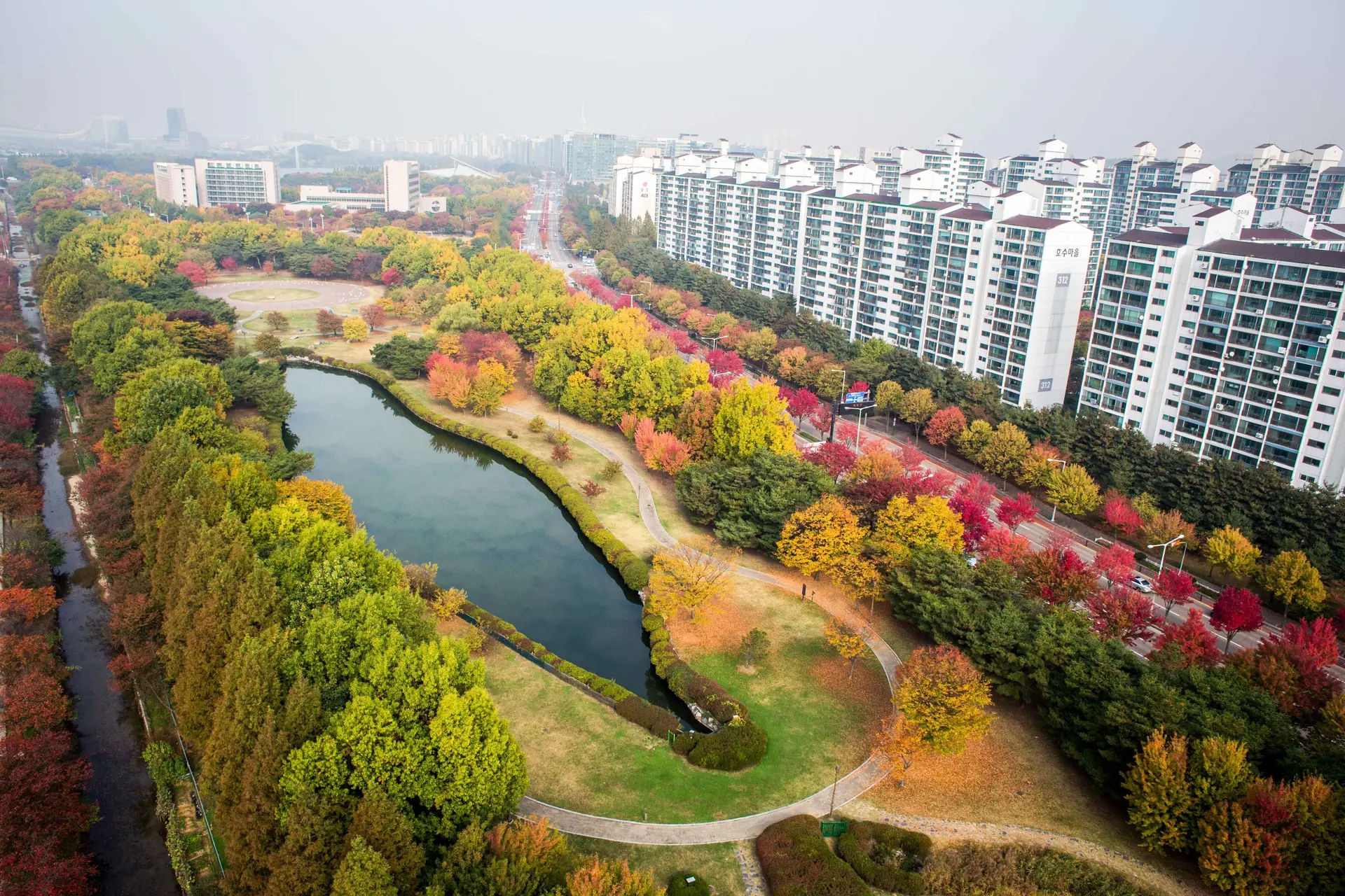 Aerial view of Ilsan Lake Park in autumn featuring vibrant red and yellow foliage, the central lake, and high-rise apartments in Goyang, South Korea.