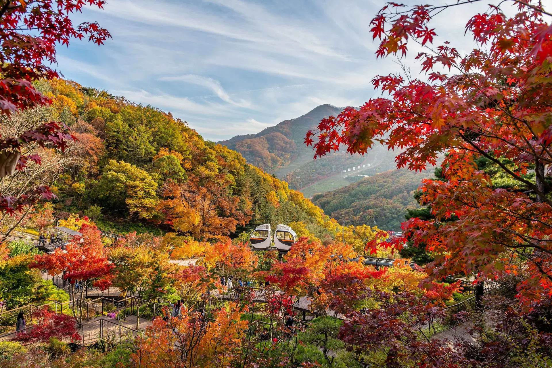 Scenic autumn view of Namhansanseong Fortress, a UNESCO World Heritage site in Gyeonggi-do, South Korea. The image features vibrant orange and yellow fall foliage surrounding the historic stone walls and gate, with hikers walking along the trail near Seoul.