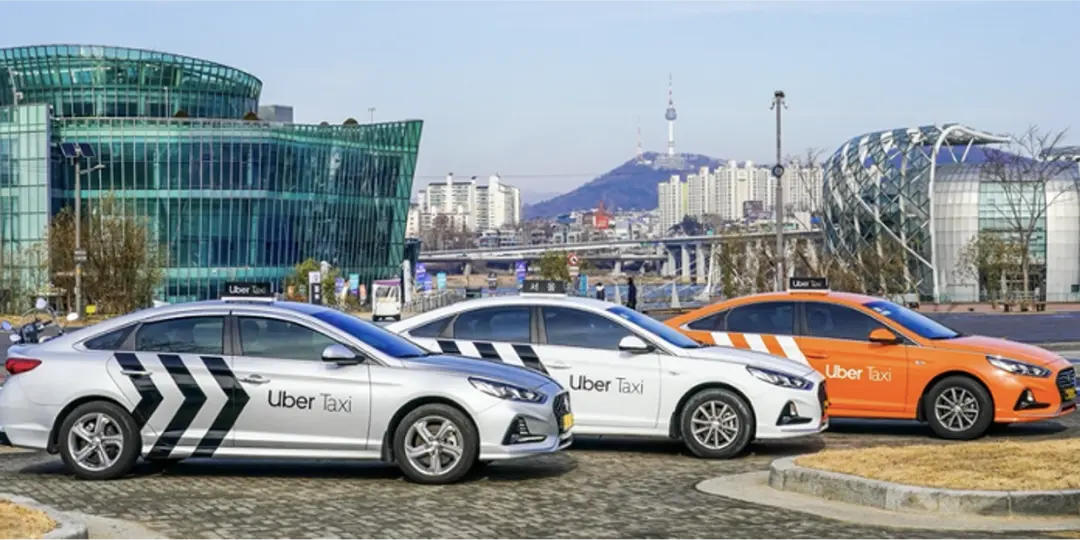 hree Uber Taxi vehicles (silver, white, and orange models) parked in a row in Seoul, South Korea, with the N Seoul Tower and city skyline visible in the background.