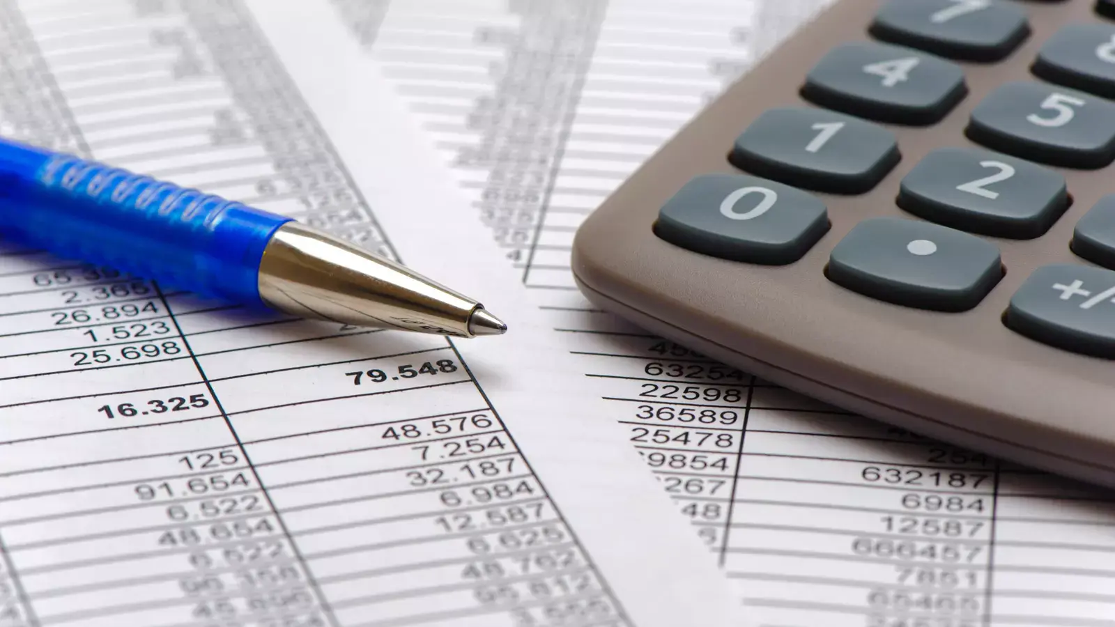 복잡한 숫자 데이터가 기록된 스프레드시트 서류, 그 위에 놓인 파란색 볼펜과 회색 계산기의 일부분