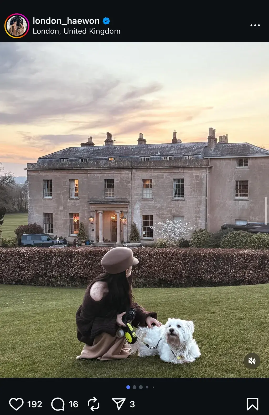 노을 지는 분홍빛 하늘 아래, 웅장한 고풍스러운 저택 앞 잔디밭에서 갈색 베레모와 스웨터를 입은 여성이 하얀 강아지와 함께 여유로운 시간을 보내고 있는 모습의 인스타그램 게시글