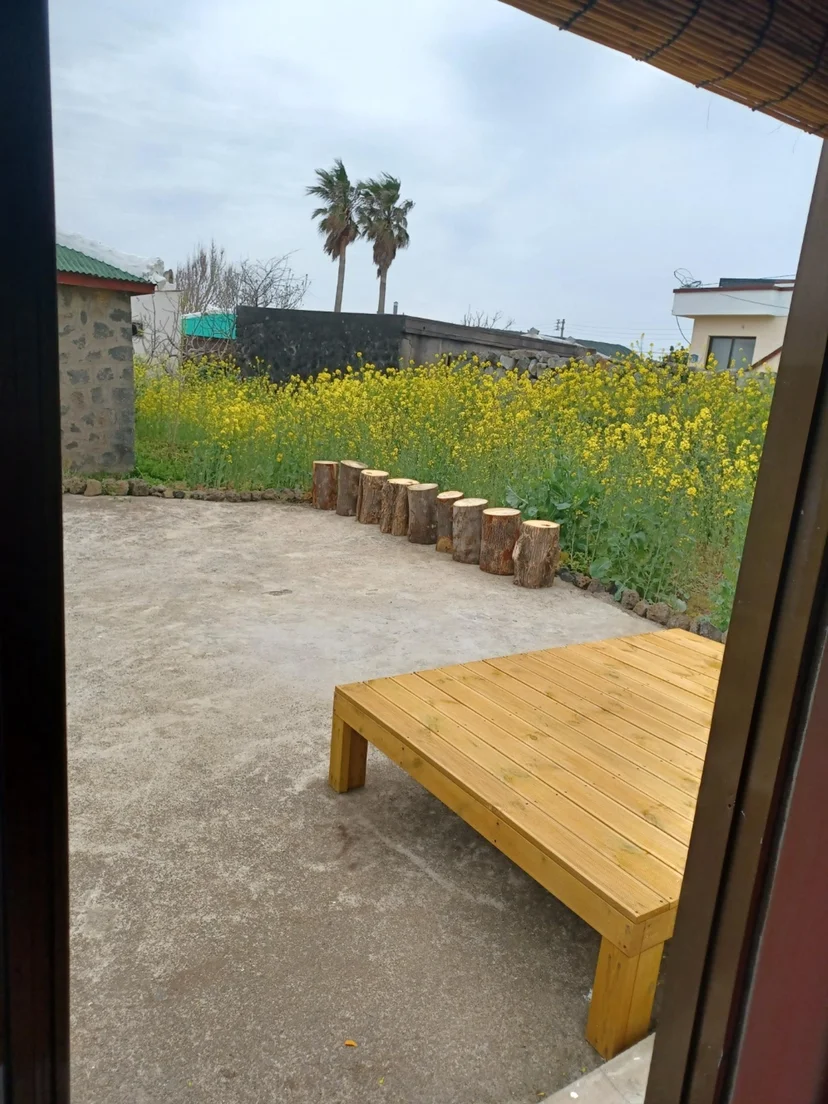 Outdoor yard with a wooden deck facing a yellow canola flower field, a palm tree, and a stone wall (Listing ID : 1954)