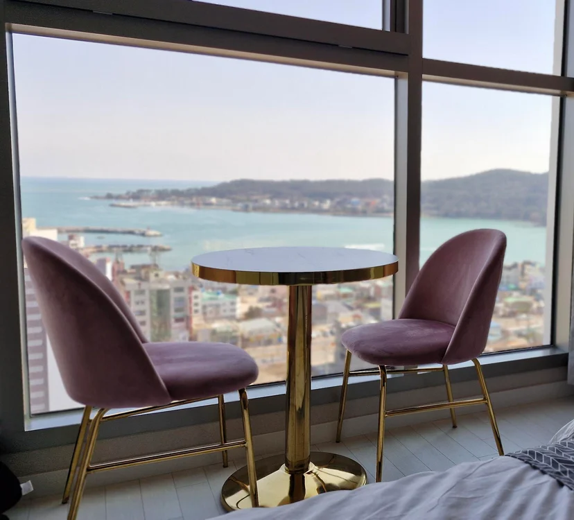 Round table and two pink velvet chairs by a window with an ocean and coastal town view (Listing ID : 22344)