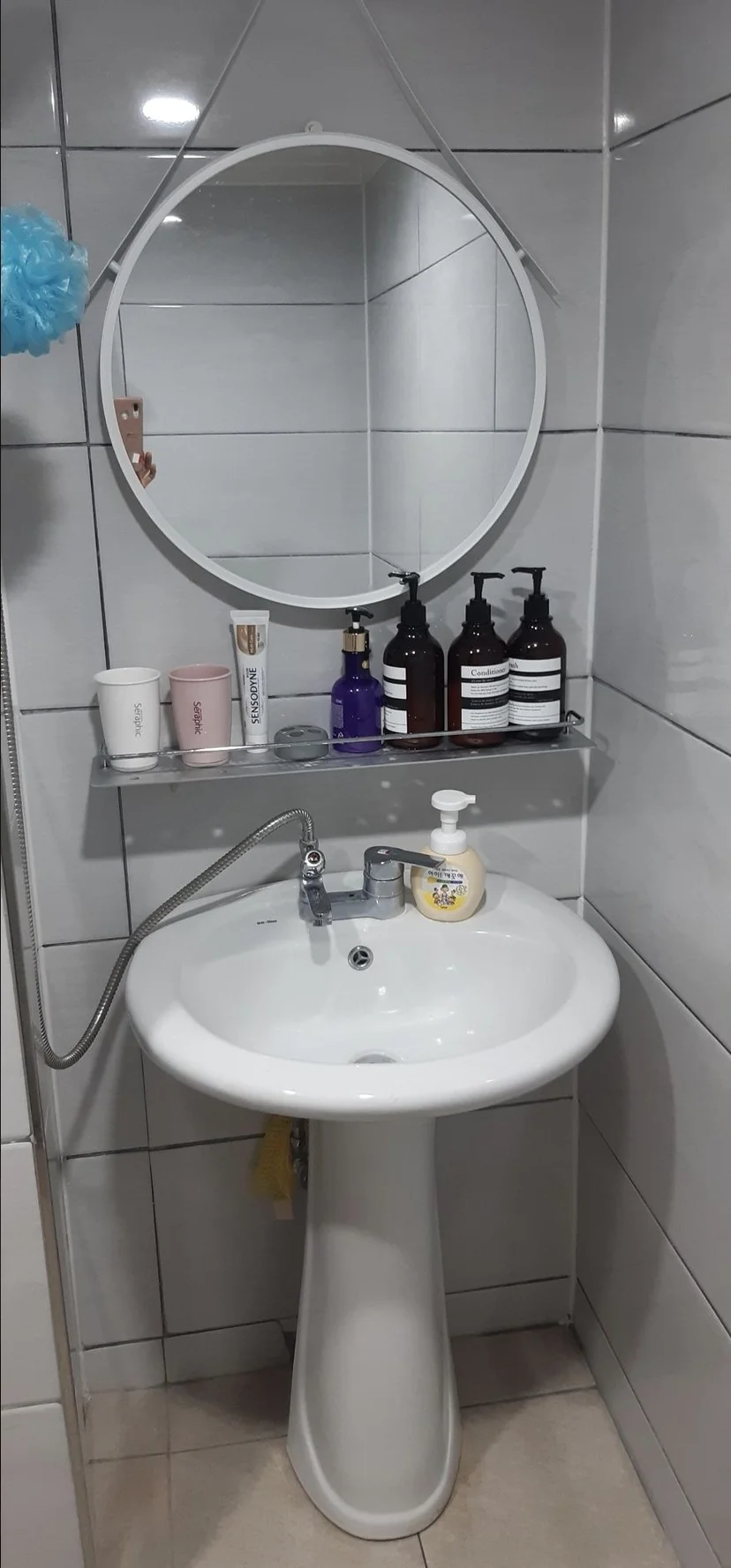 Clean tiled bathroom with a white pedestal sink, chrome faucet, round mirror, and toiletry shelf (Listing ID : 1954)