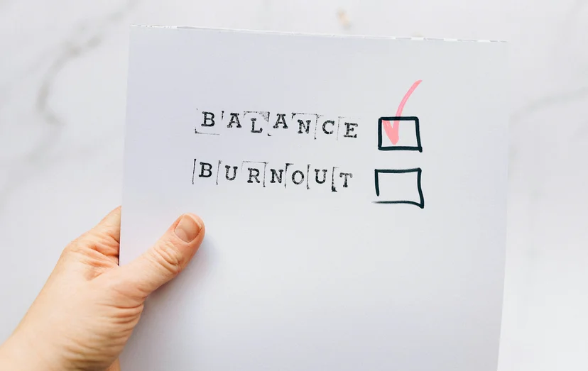 하얀 A4용지를 들고 있는 손. A4용지에는 영단어 'BALANCE'와 'BURNOUT'이 쓰여 있고, 각각의 단어 옆에 체크박스가 있다. 'BALANCE'단어 옆에만 체크 표시가 되어 있어 직무 소진 방지를 의미하는 이미지이다.