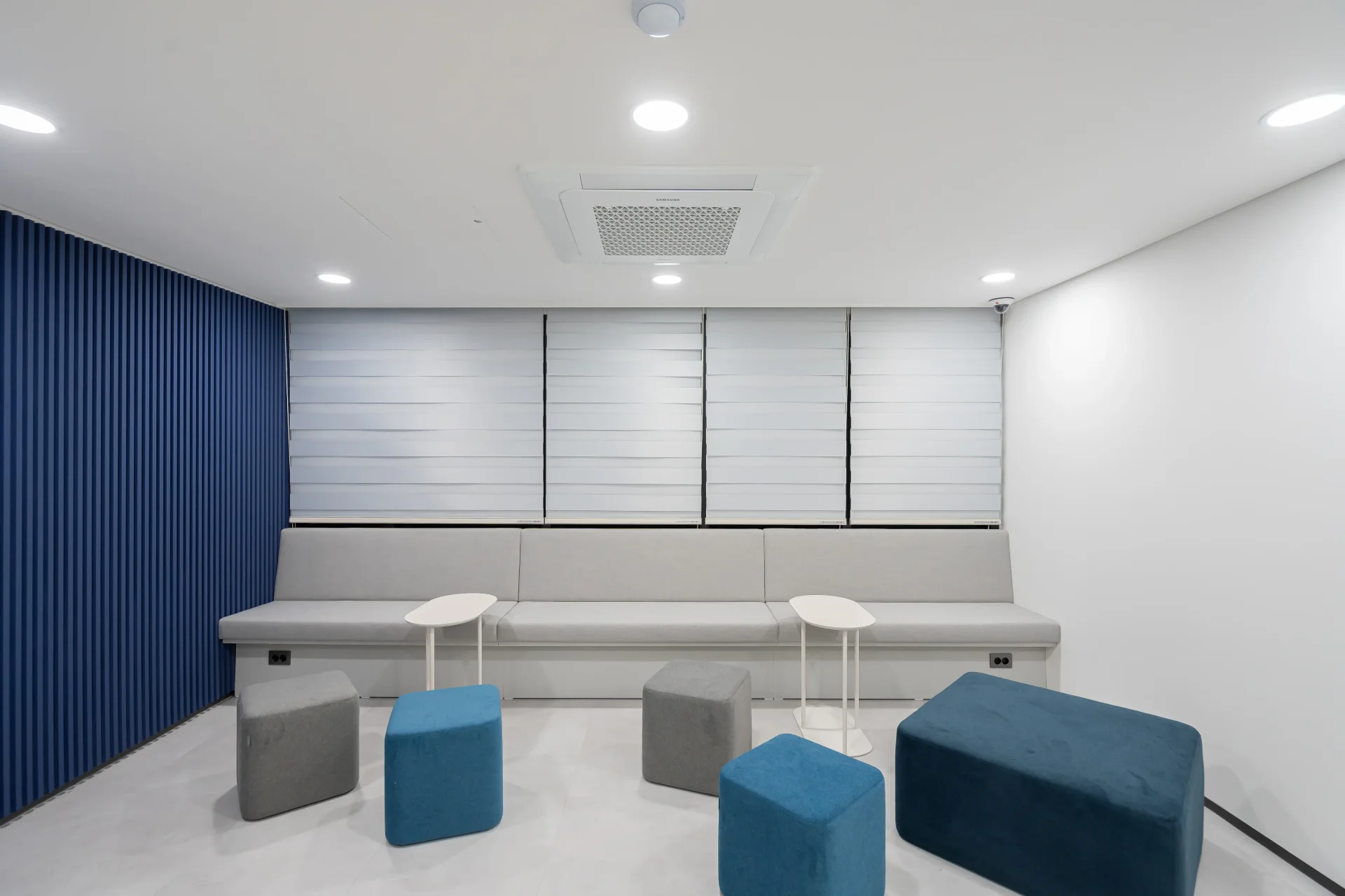 미니멀한 디자인의 라운지 공간 인테리어로, 블루와 그레이 컬러의 좌식 큐브형 의자와 흰색 벤치 소파가 조화롭게 배치되어 있습니다. 창가에 위치한 휴게 공간으로 현대적인 오피스 인테리어 디자인을 보여줍니다.