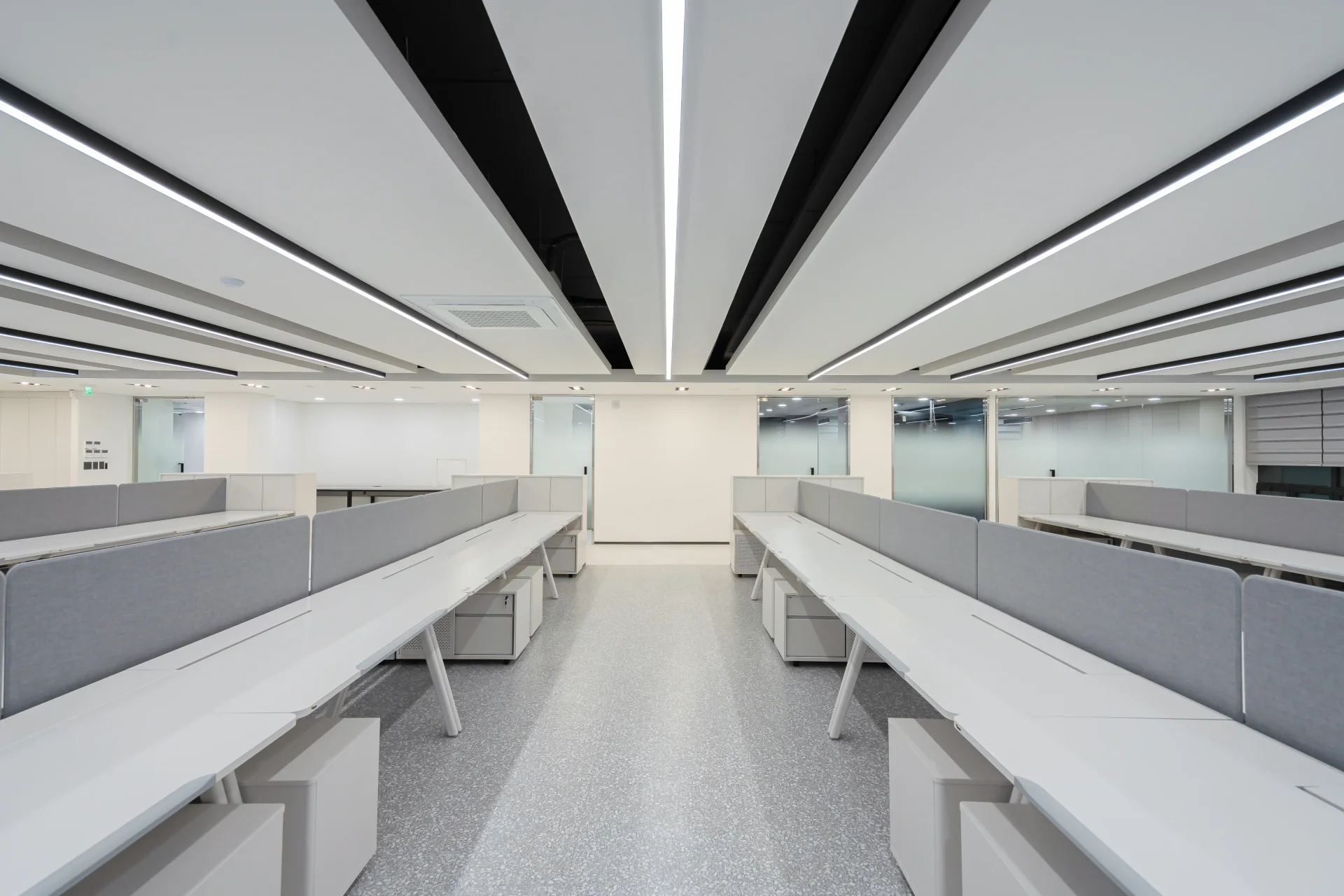 개방형 오피스 인테리어의 워크스테이션으로, 흰색 긴 테이블과 회색 분리 패널로 구성되어 있습니다. 천장의 선형 조명과 깔끔한 바닥이 특징적인 현대적 사무실 인테리어로 효율적인 업무 환경을 제공합니다.