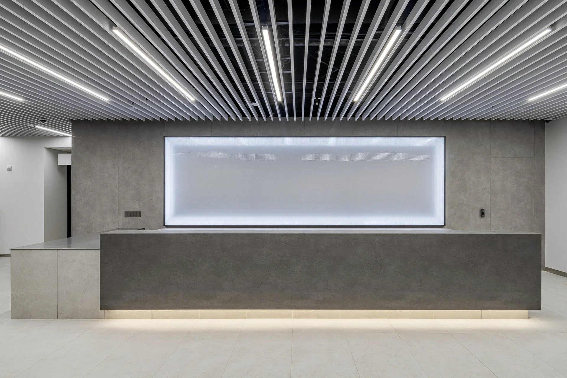 미니멀한 디자인의 리셉션 데스크 인테리어로, 콘크리트 마감과 백라이트 패널이 특징적입니다. 천장의 바 형태 조명 구조물이 독특한 분위기를 연출하는 현대적인 오피스 로비 인테리어입니다.