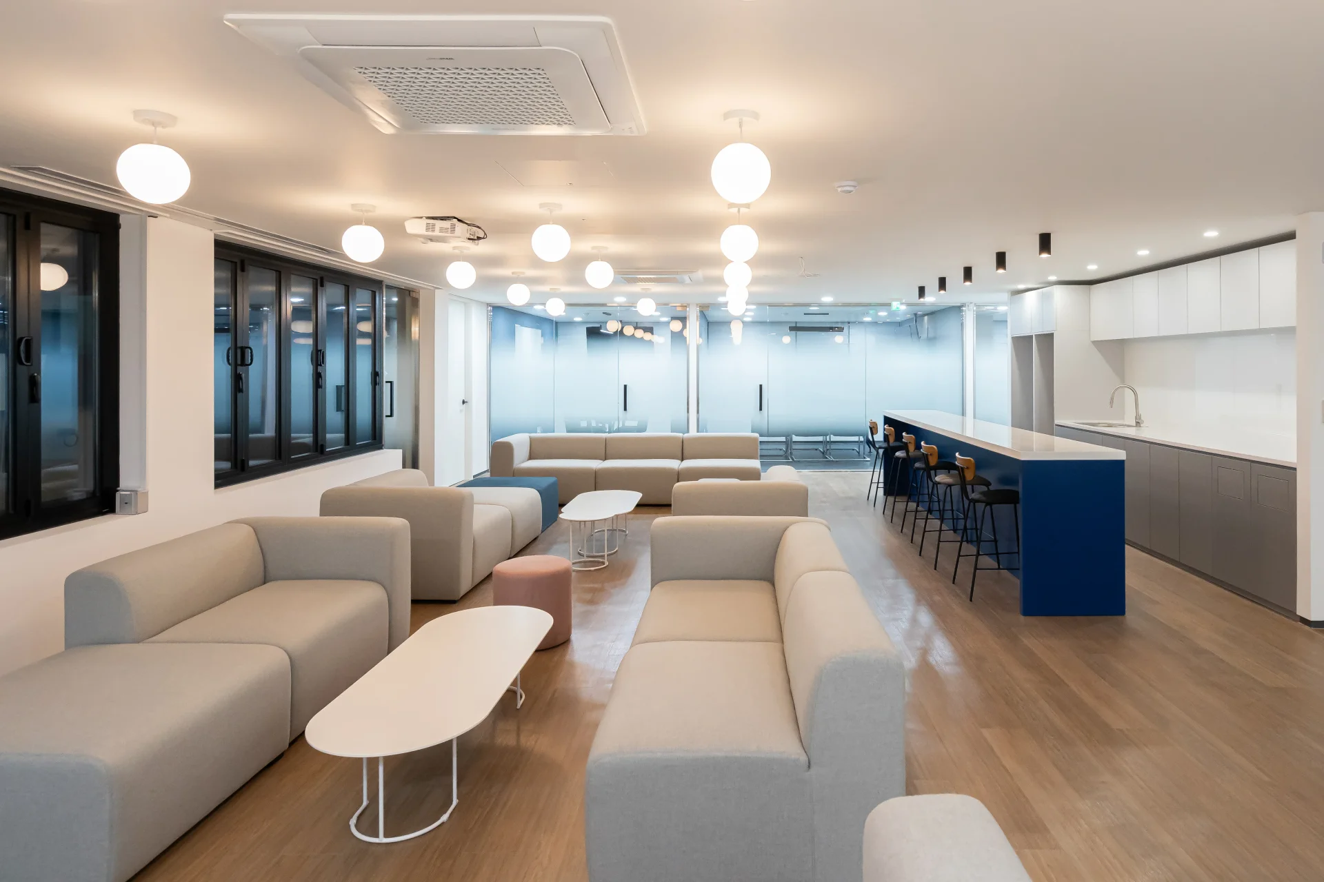 현대적인 캔틴 겸 라운지 공간 인테리어로, 베이지색 모듈형 소파와 블루 컬러 주방 카운터가 조화를 이룹니다. 원형 펜던트 조명과 목재 바닥으로 따뜻한 분위기를 연출한 스타트업 사옥 인테리어입니다.