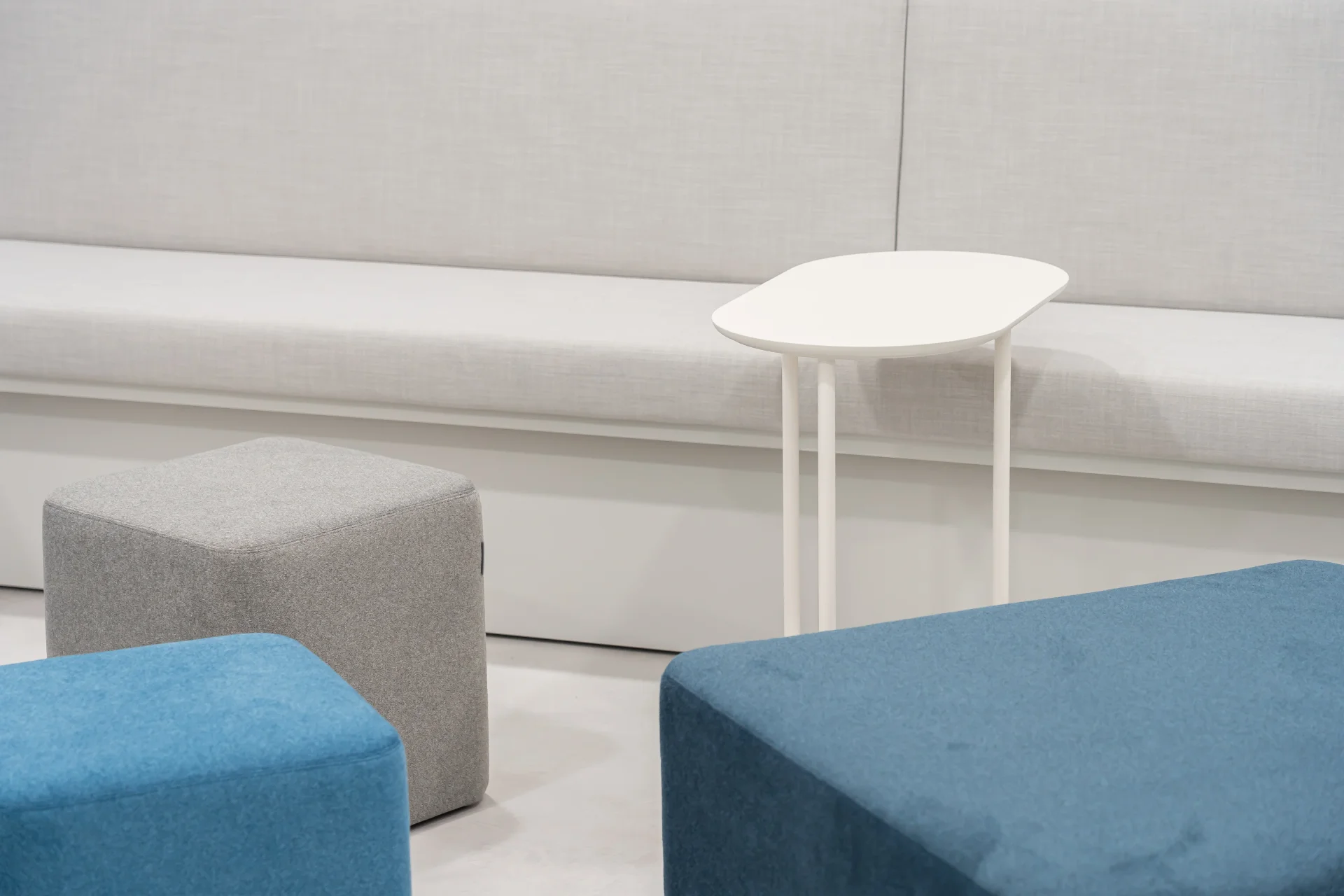 현대적인 오피스 인테리어의 휴게 공간으로, 청록색과 회색 톤의 사각 패브릭 좌식 큐브가 배치되어 있고 흰색의 간결한 사이드 테이블이 함께 어우러져 있습니다. 벽면에는 깔끔한 흰색 벤치형 소파가 설치되어 있어 직원들의 캐주얼한 미팅이나 휴식에 적합한 스타트업 사무실 인테리어 디자인을 보여줍니다.