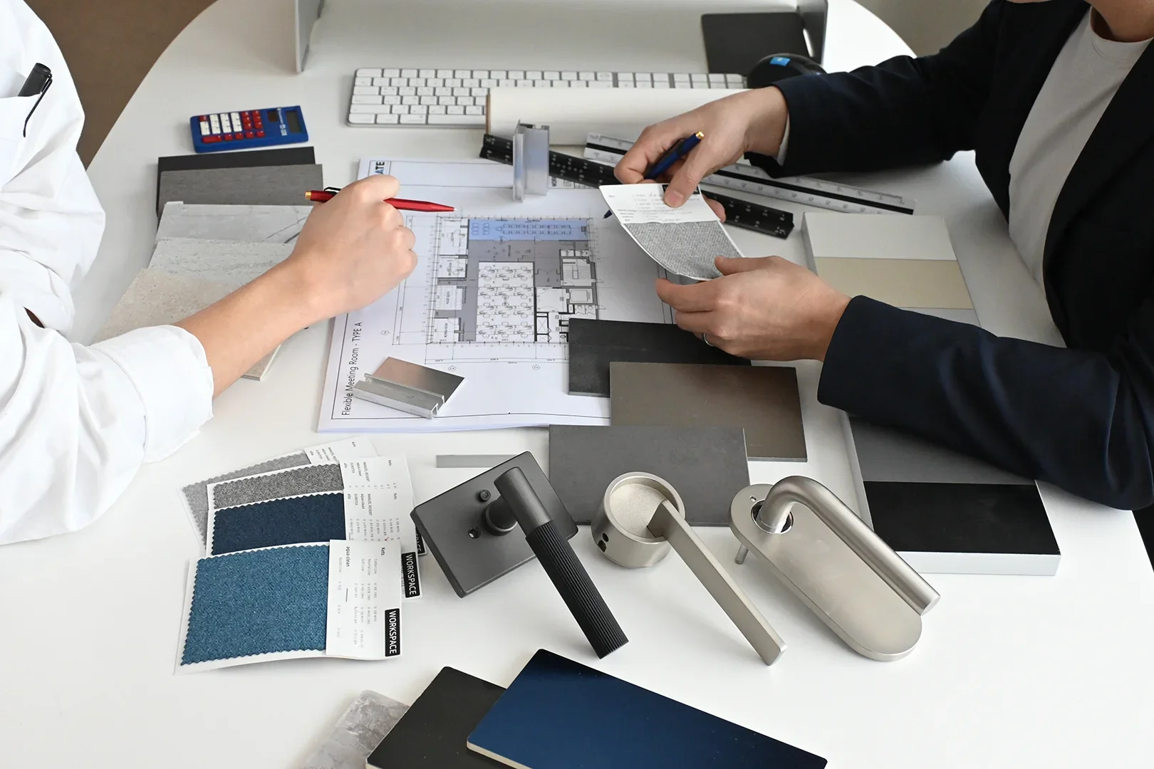 사무실 인테리어 디자인 미팅 장면으로, 두 명의 전문가가 테이블에서 도면과 소재 샘플을 검토하고 있습니다. 흰색 의상을 입은 디자이너가 빨간 펜으로 평면도를 가리키고 있으며, 검은색 의상을 입은 담당자가 소재 샘플을 살펴보고 있습니다. 테이블 위에는 파란색 패브릭 샘플, 그레이 톤의 마감재 견본, 문손잡이와 같은 하드웨어 샘플들이 배치되어 있습니다. 사옥 인테리어 프로젝트의 디자인 단계에서 소재와 색상을 선정하는 전문적인 협업 과정을 보여주는 장면입니다.