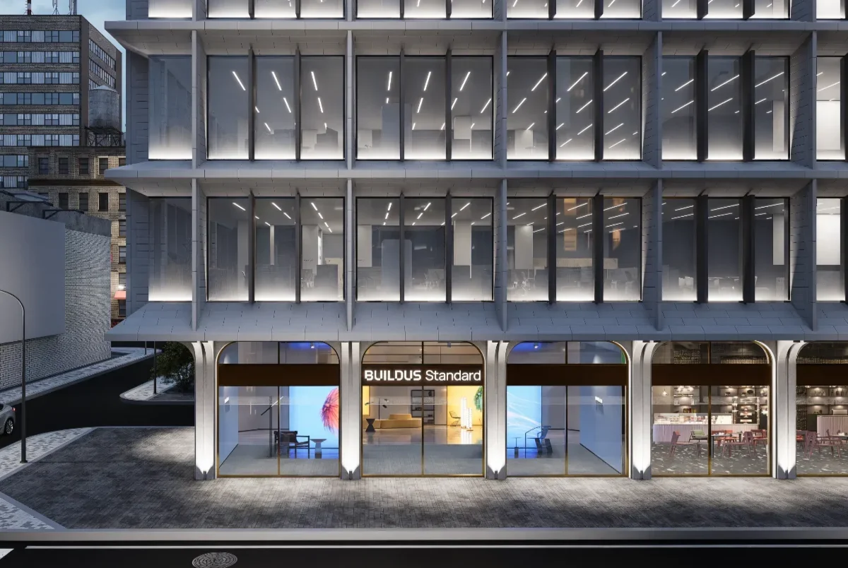현대적인 디자인의 BUILDUS Standard 오피스 빌딩 외관으로, 야간에 촬영된 모습입니다. 3층 구조의 건물로, 대형 유리창과 세련된 조명이 특징적입니다. 1층은 "BUILDUS Standard"라는 간판이 빛나는 쇼룸형 전시 공간으로 구성되어 있으며, 골드 프레임의 유리 출입구가 고급스럽습니다. 상층부는 선형 조명이 설치된 오피스 공간으로, 전체적으로 미니멀하고 세련된 디자인의 사옥 인테리어와 외관이 조화를 이루고 있습니다. 전문적인 사무실 인테리어 업체의 아이덴티티가 건물 디자인에 잘 반영되어 있습니다.