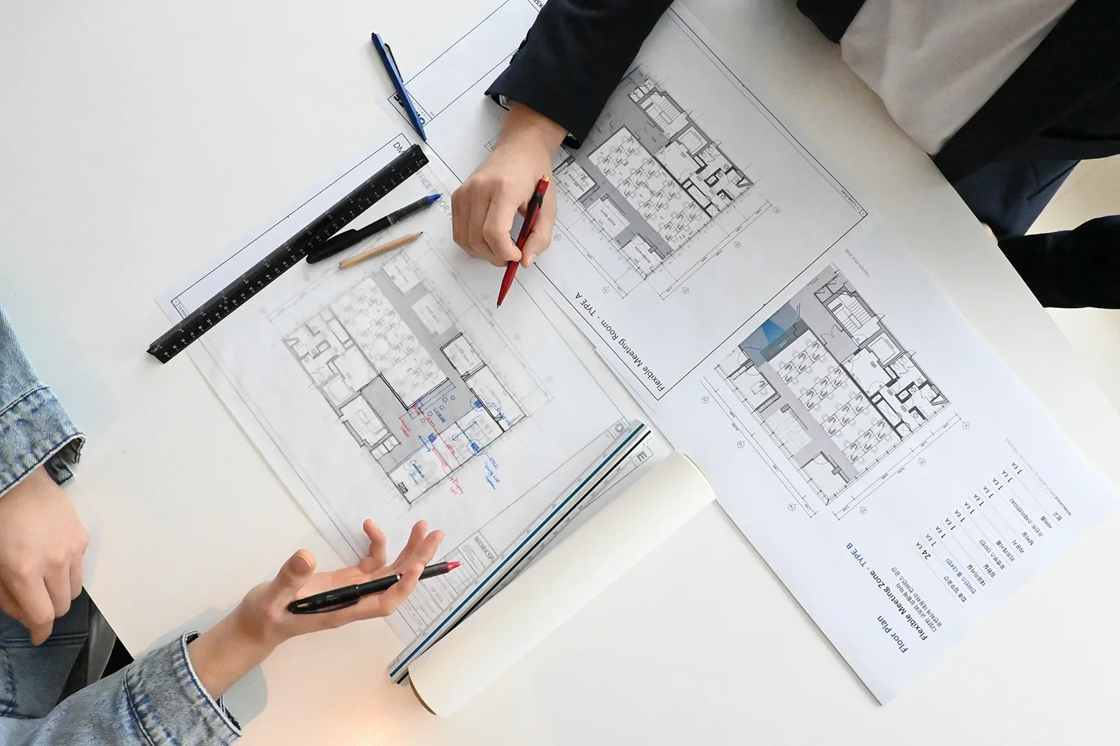 사무실 인테리어 설계 미팅 장면으로, 흰색 테이블 위에 여러 장의 건축 평면도가 펼쳐져 있습니다. 두 명의 전문가가 도면을 검토하며 논의하고 있으며, 한 사람은 빨간색 펜으로 특정 부분을 가리키고 있고 다른 사람은 펜을 들고 의견을 제시하고 있습니다. 
