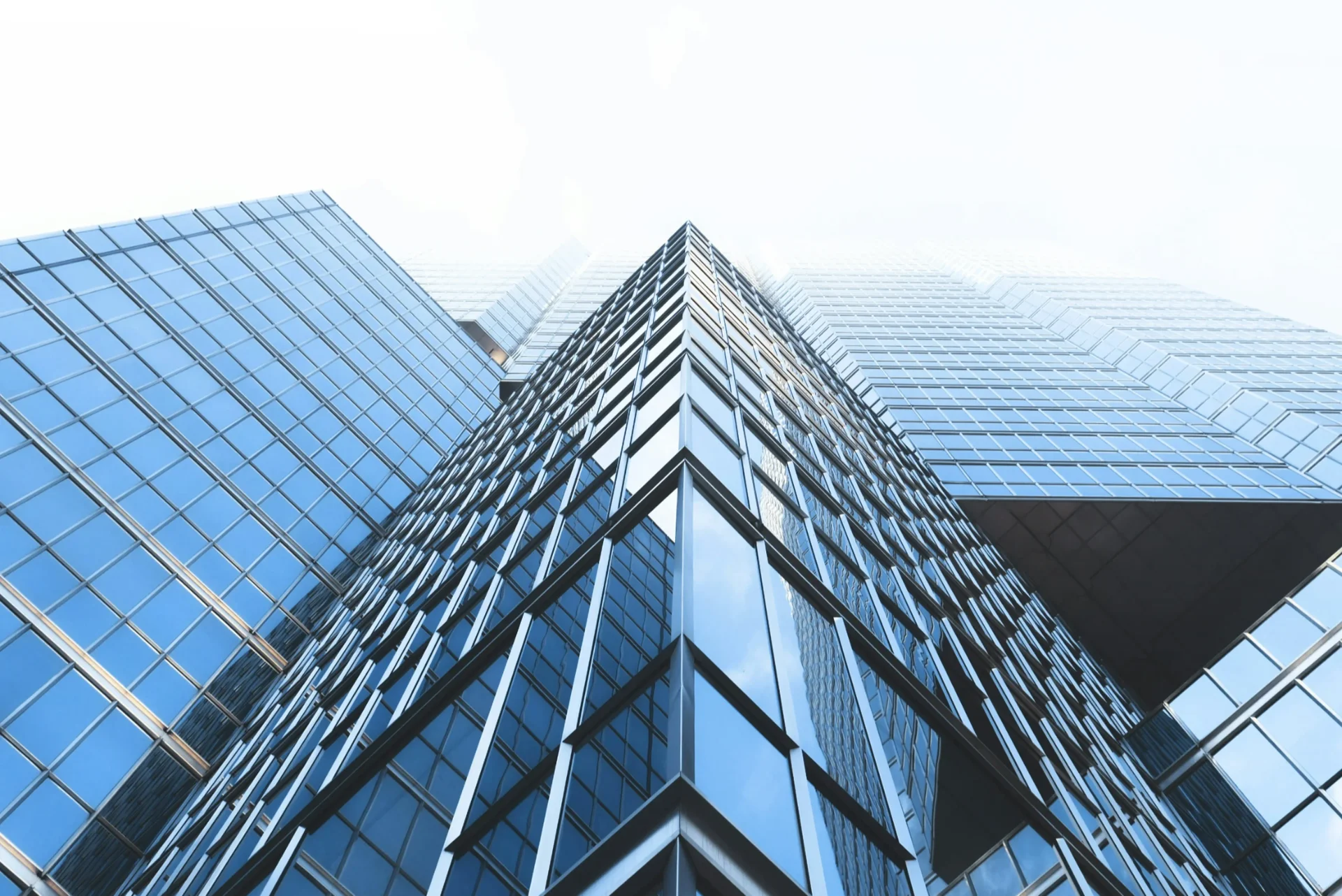현대적인 오피스 빌딩의 외관을 아래에서 위로 올려다 본 모습입니다. 전체적으로 푸른색 유리 커튼월로 이루어진 고층 건물로, 각진 모서리 부분이 강조되어 있습니다. 맑은 하늘을 배경으로 건물의 유리 외벽이 반사되어 세련되고 전문적인 느낌을 주는 사무실 인테리어와 사옥 건축의 현대적 트렌드를 보여줍니다. 