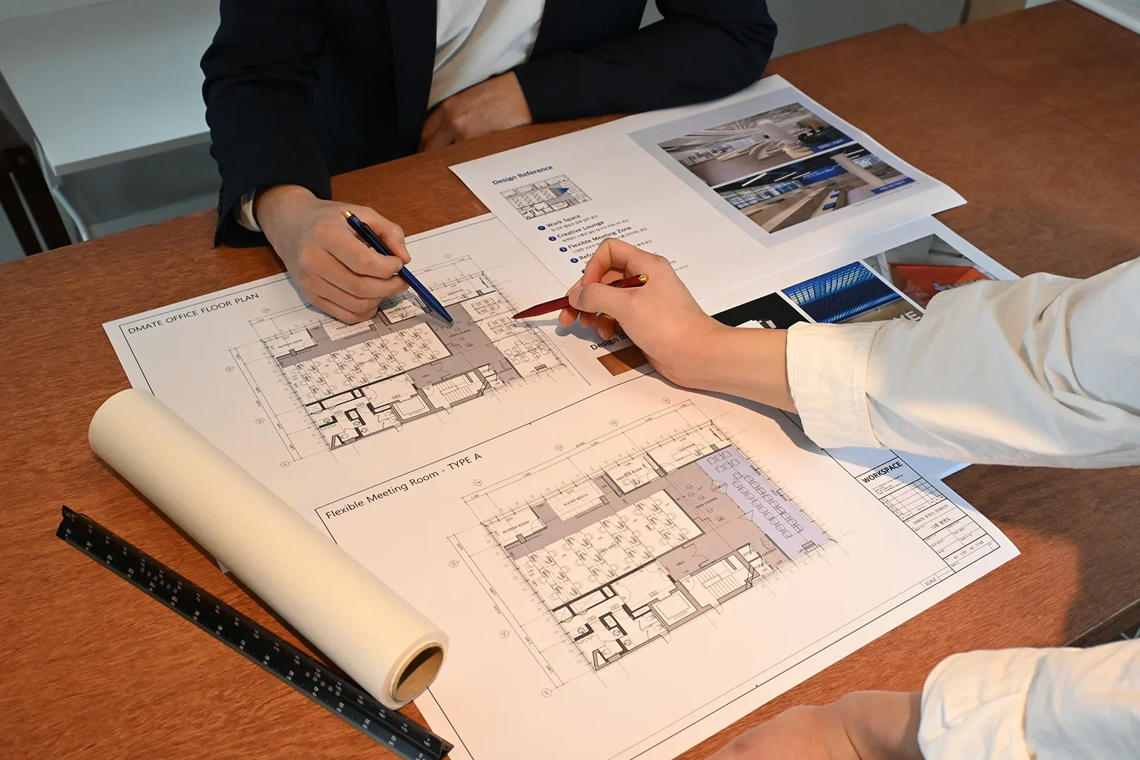 사무실 인테리어 컨설팅 현장에서 두 명의 전문가가 오피스 평면도를 검토하는 모습입니다. 목재 테이블 위에는 "OFFICE FLOOR PLAN"이라고 표시된 상세 도면과 "Flexible Meeting Room - TYPE A" 영역이 표시된 설계도면이 펼쳐져 있으며, 한 사람은 네이비 재킷을, 다른 사람은 흰색 셔츠를 입고 있습니다. 청사진 근처에는 설계 참고 자료와 오피스 인테리어 사례 사진이 담긴 문서가 있고, 자와 도면 롤이 함께 놓여있습니다. 두 전문가는 펜으로 도면의 특정 부분을 가리키며 사옥 인테리어 계획에 대해 논의하고 있습니다. 이는 전문적인 사무실 공간 설계 과정의 중요한 컨설팅 단계를 보여주는 장면입니다.