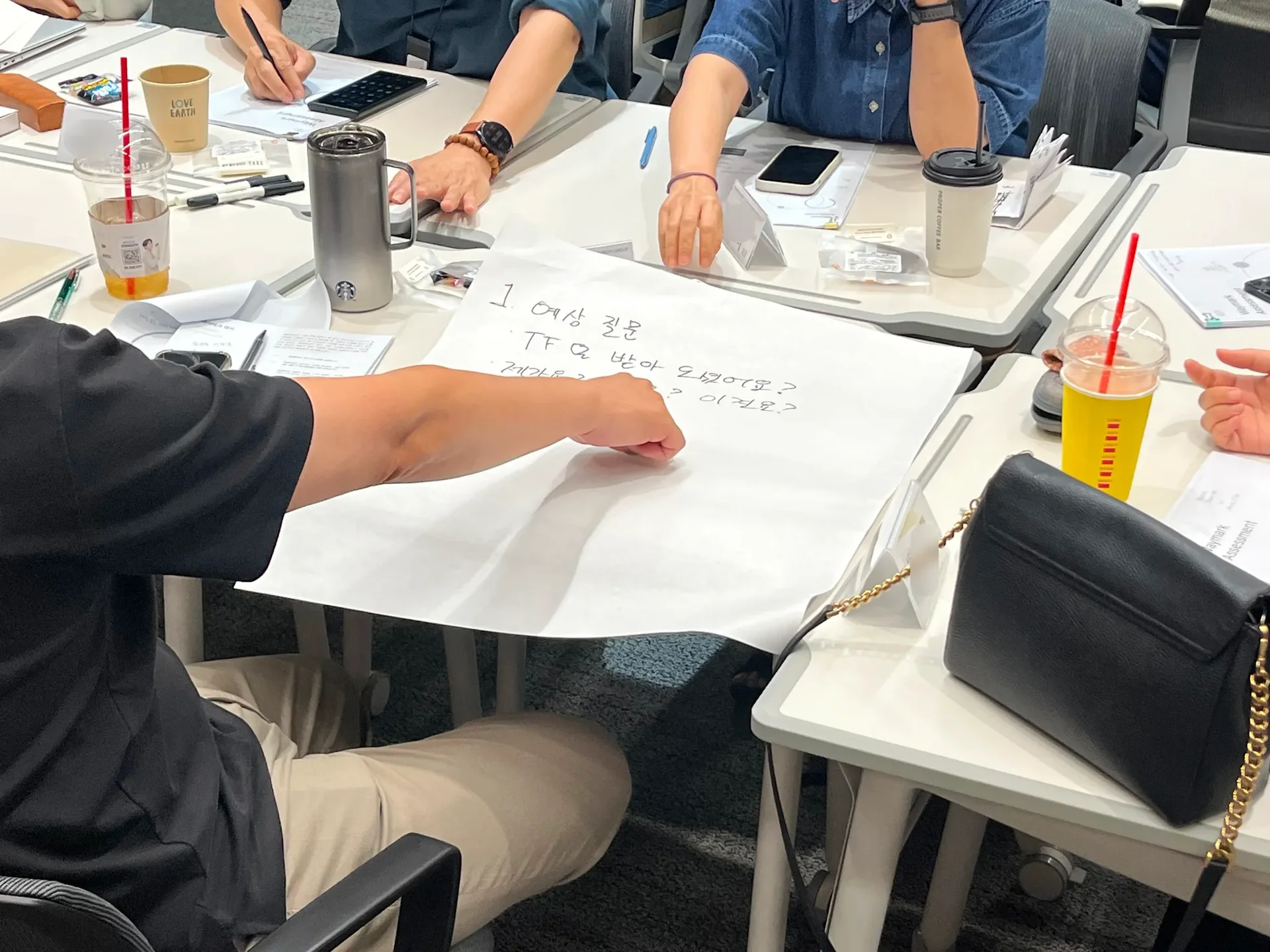 테이블에 놓인 흰 전지 위에 큰 글씨로 토의 내용이 적혀있다. 한 남성이 전지에 적힌 글씨를 가리키고 있다.