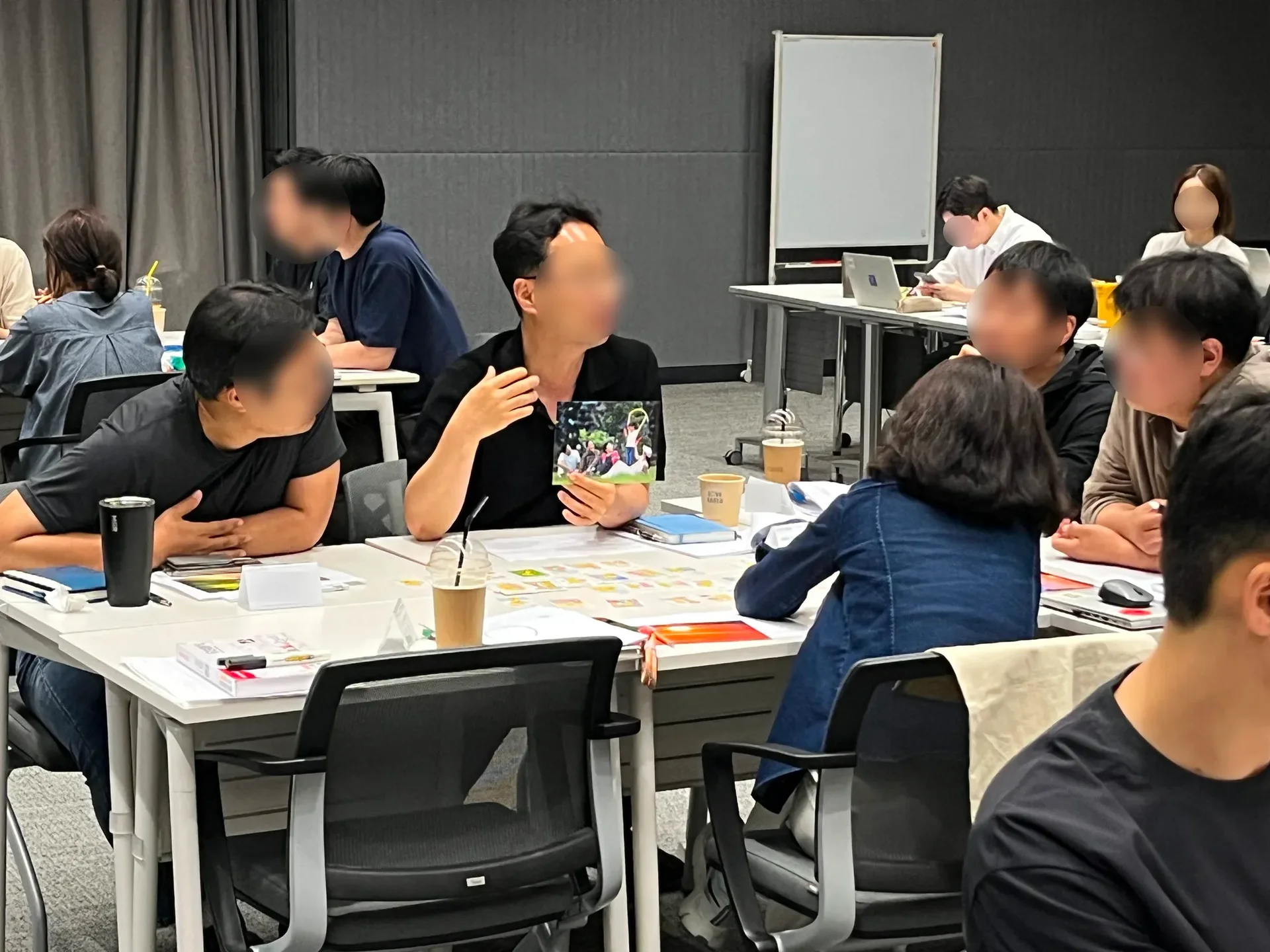 한 남성이 사진을 들고 손으로 제스처를 취하며 자신의 지향점을 설명하고 있다. 다른 조원들은 사진과 해당 남성을 보며 경청하고 있다.