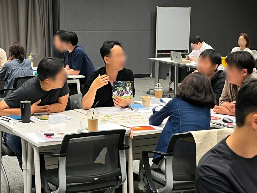 한 남성이 사진을 들고 손으로 제스처를 취하며 자신의 지향점을 설명하고 있다. 다른 조원들은 사진과 해당 남성을 보며 경청하고 있다.