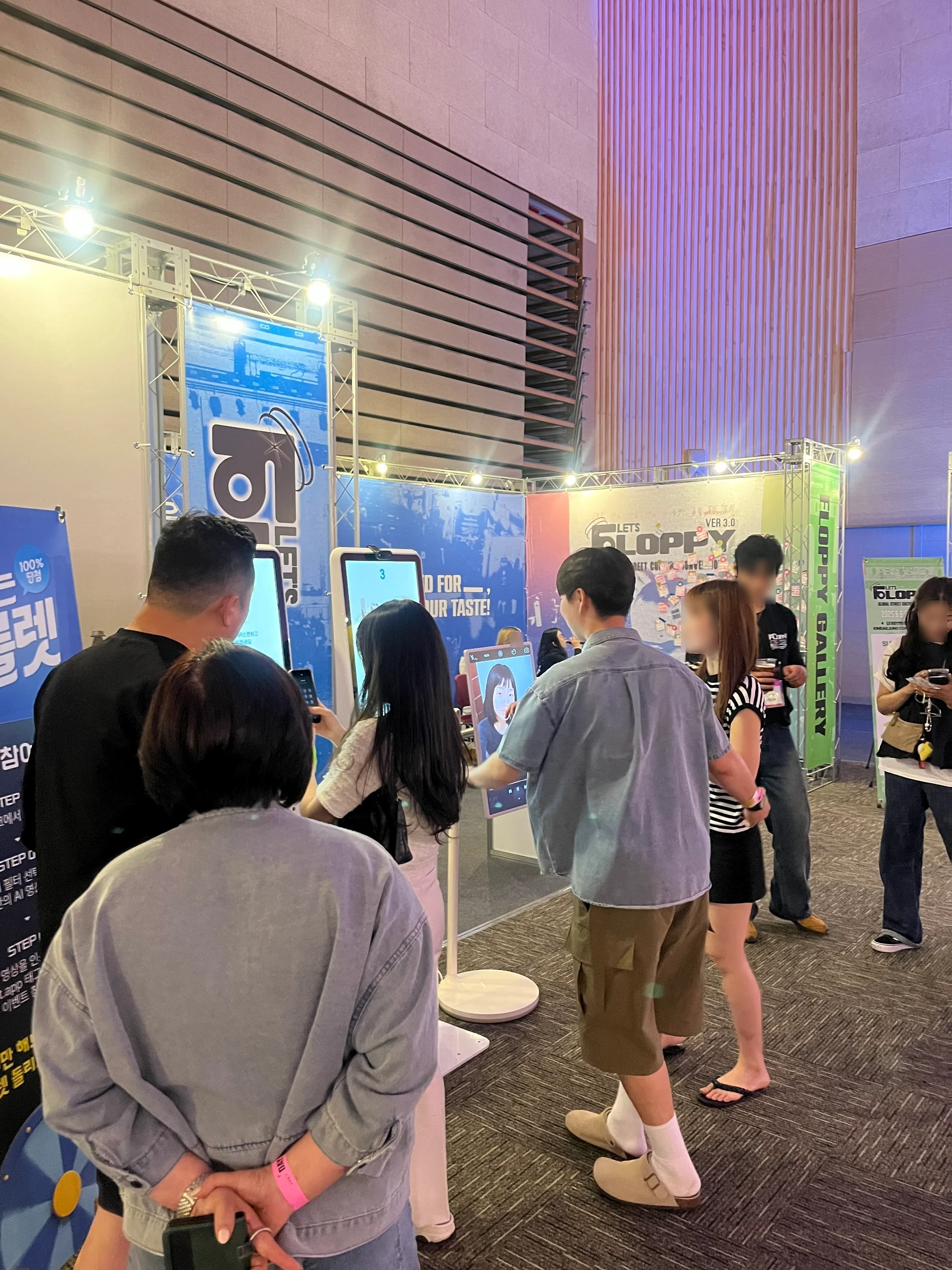 실내 전시 공간에서 여러 명의 관람객들이 키오스크 앞에 줄을 서 있거나 체험 중이다. 키오스크 화면에는 캐릭터 이미지가 표시되어 있고, 참가자들은 직접 입력하거나 촬영 중인 모습이다. 부스 상단에는 ‘플로피(Floppy)’ 로고와 홍보 배너가 설치되어 있으며, 부스 주변은 조명으로 밝게 조성되어 있다. 대부분의 참가자들이 편안한 복장으로 관람 중이며, 배경에는 목재 패널 벽과 전시용 구조물이 설치되어 있다.