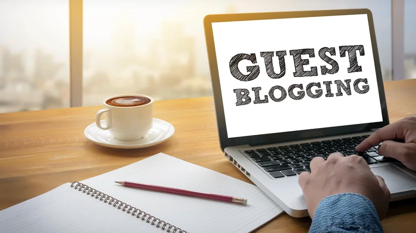 Person typing on a laptop with 'Guest Blogging' on the screen, next to a coffee cup and notepad on a wooden desk