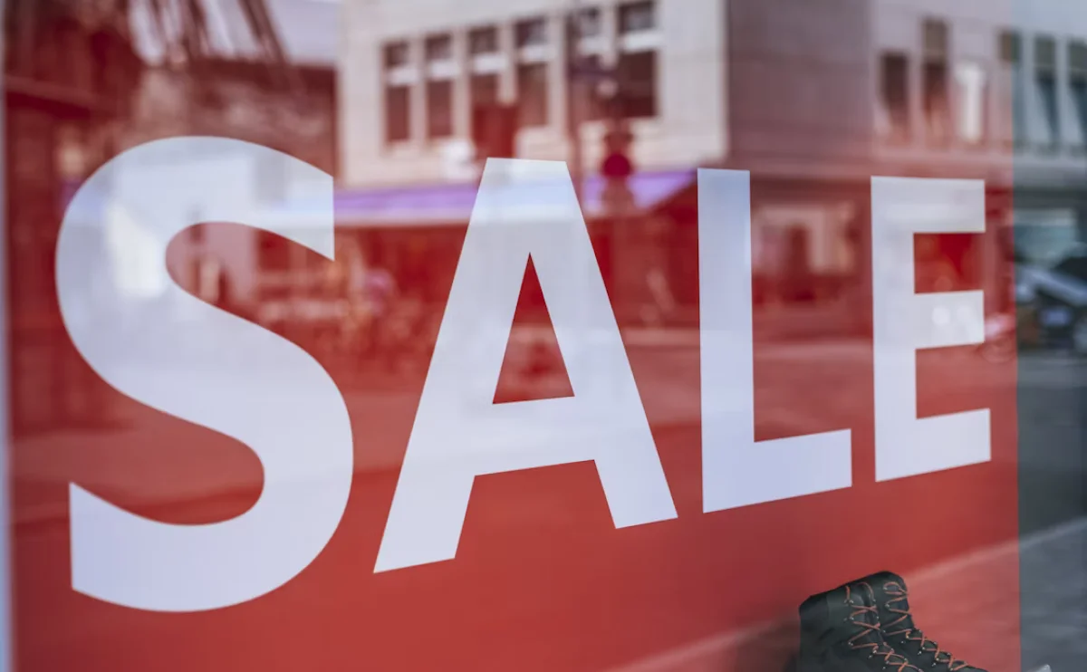 쇼윈도에 크게 적힌 'SALE' 문구가 보이는 이미지. 배경에는 도시 거리와 건물이 비쳐 보이며, 오른쪽 하단에는 검은색 등산화 한 켤레가 일부 보입니다
