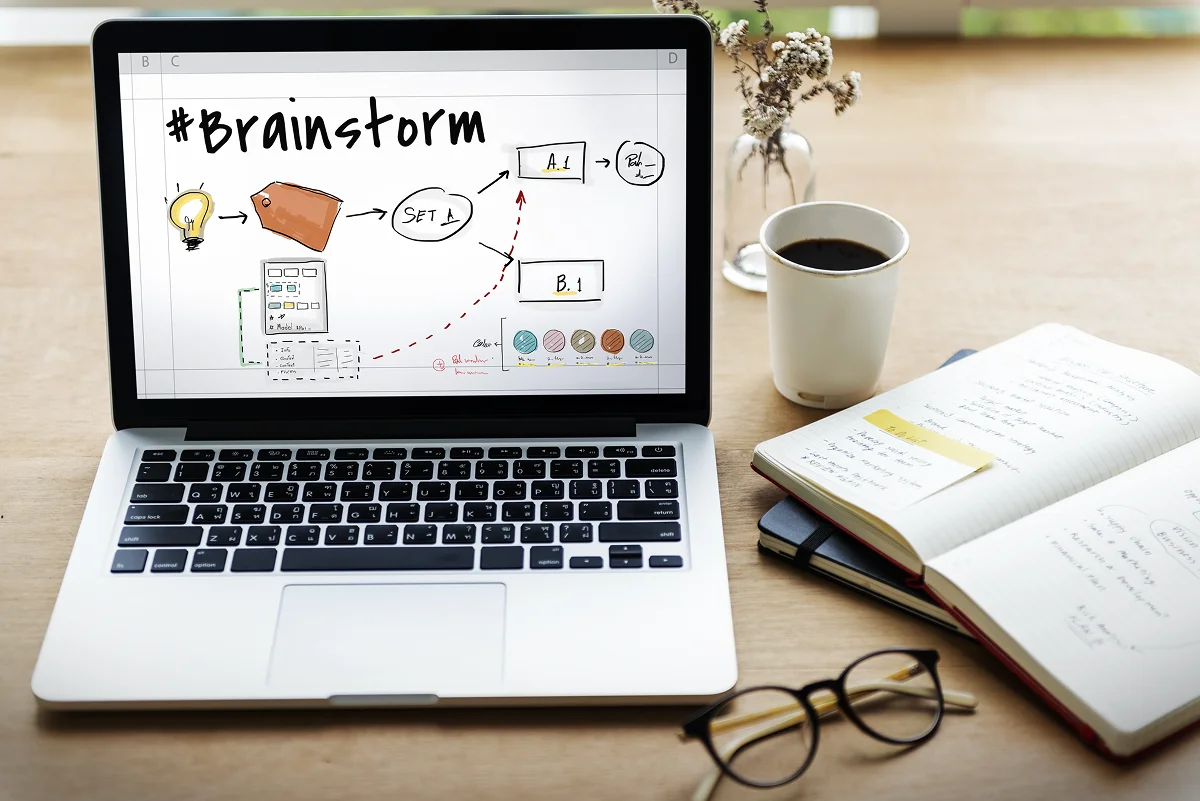 #Brainstorm’이라는 단어와 아이디어 흐름도가 표시된 노트북 화면. 노트북 옆에는 커피컵, 손글씨가 적힌 노트, 안경, 연필, 작은 꽃병이 나무 테이블 위에 놓여 있는 모습