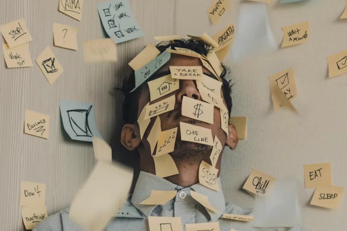 피곤한 표정의 남성이 벽에 기대어 앉아 얼굴과 몸에 수많은 포스트잇을 붙이고 있는 모습. 포스트잇에는 'Take a break', 'STOP', 'OFFLINE', 'Sleep', '$' 등의 낙서나 문구가 적혀 있으며, 혼란스러운 정신 상태와 과로, 스트레스를 시각적으로 표현하고 있다.
