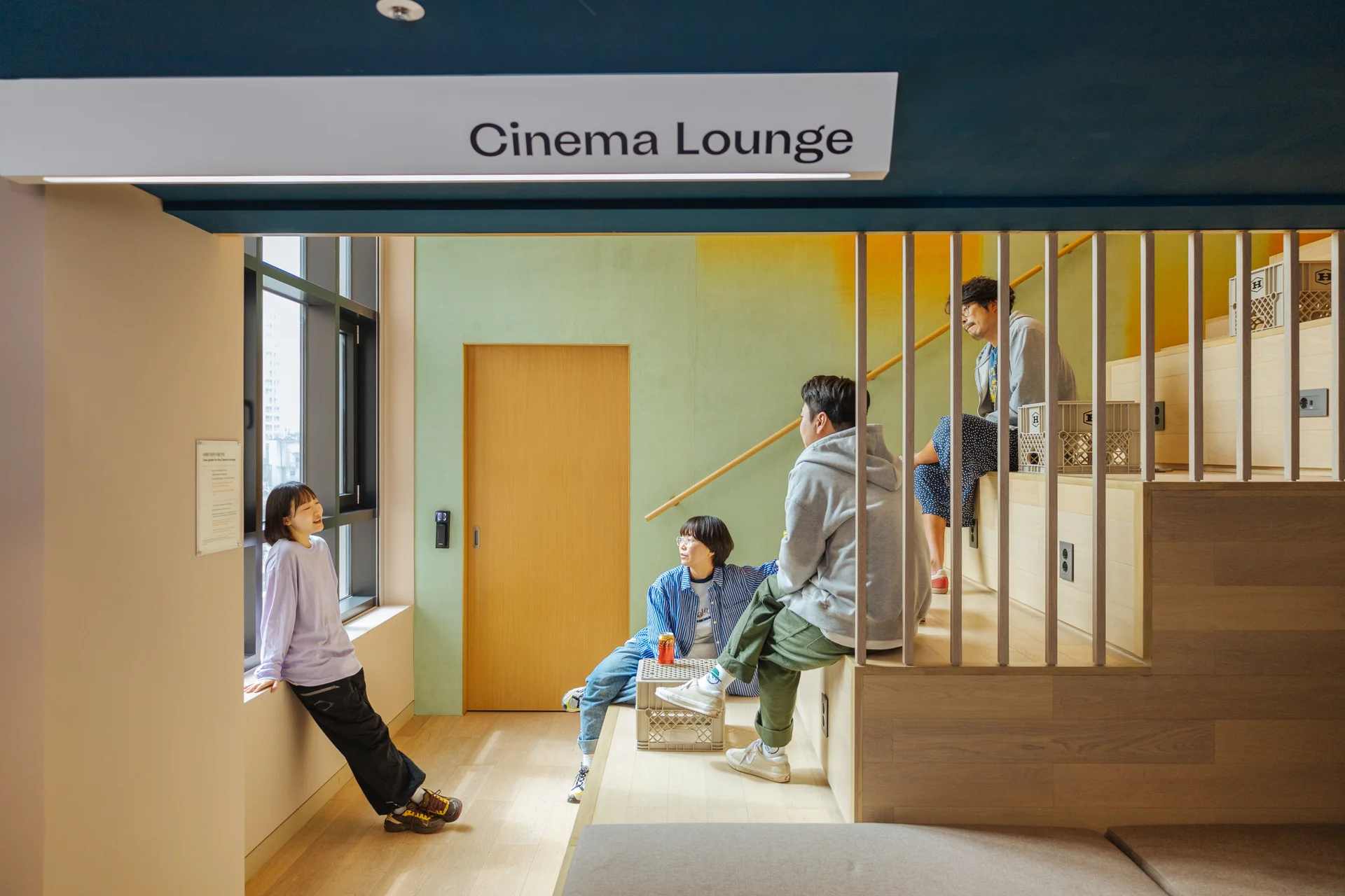 맹그로브 신촌의 시네마 라운지. 계단형 좌석이 있는 아늑한 공간에서 네 명의 사람들이 모여 대화하며 휴식을 취하고 있다. 한 사람은 창가에 기대어 서 있고, 나머지는 계단형 좌석에 앉아 서로 이야기하는 모습이다. 공간은 따뜻한 나무 소재와 파스텔 톤 벽으로 꾸며져 있으며, 라운지 상단에는 'Cinema Lounge'라는 안내판이 걸려 있다.