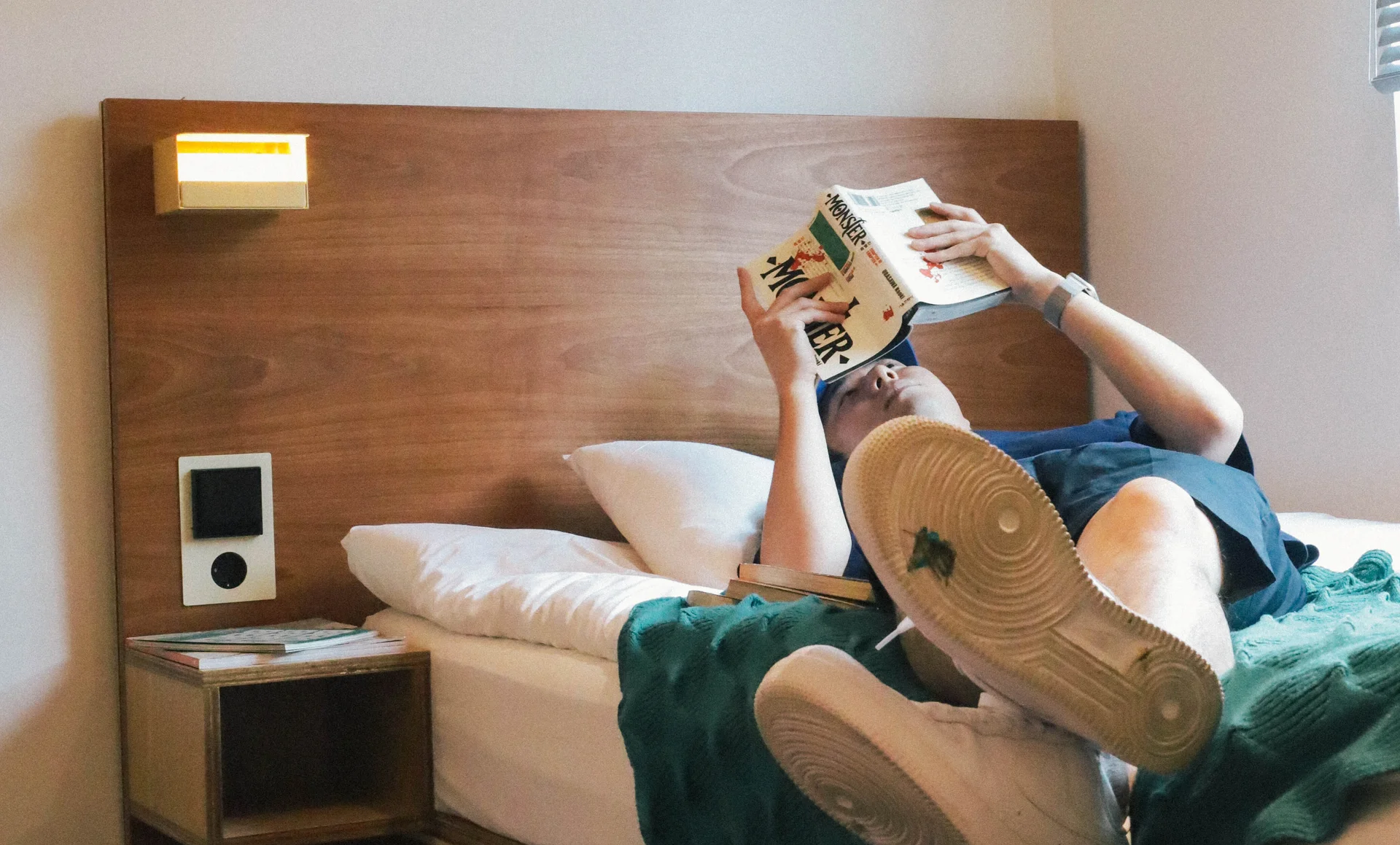 맹그로브 신설 싱글룸에서 한 사람이 침대에 누워 책을 읽고 있는 모습. 침대 옆 협탁에는 책이 놓여 있고, 따뜻한 조명이 켜져 있으며, 단정한 싱글 침대와 원목 헤드보드가 보인다.