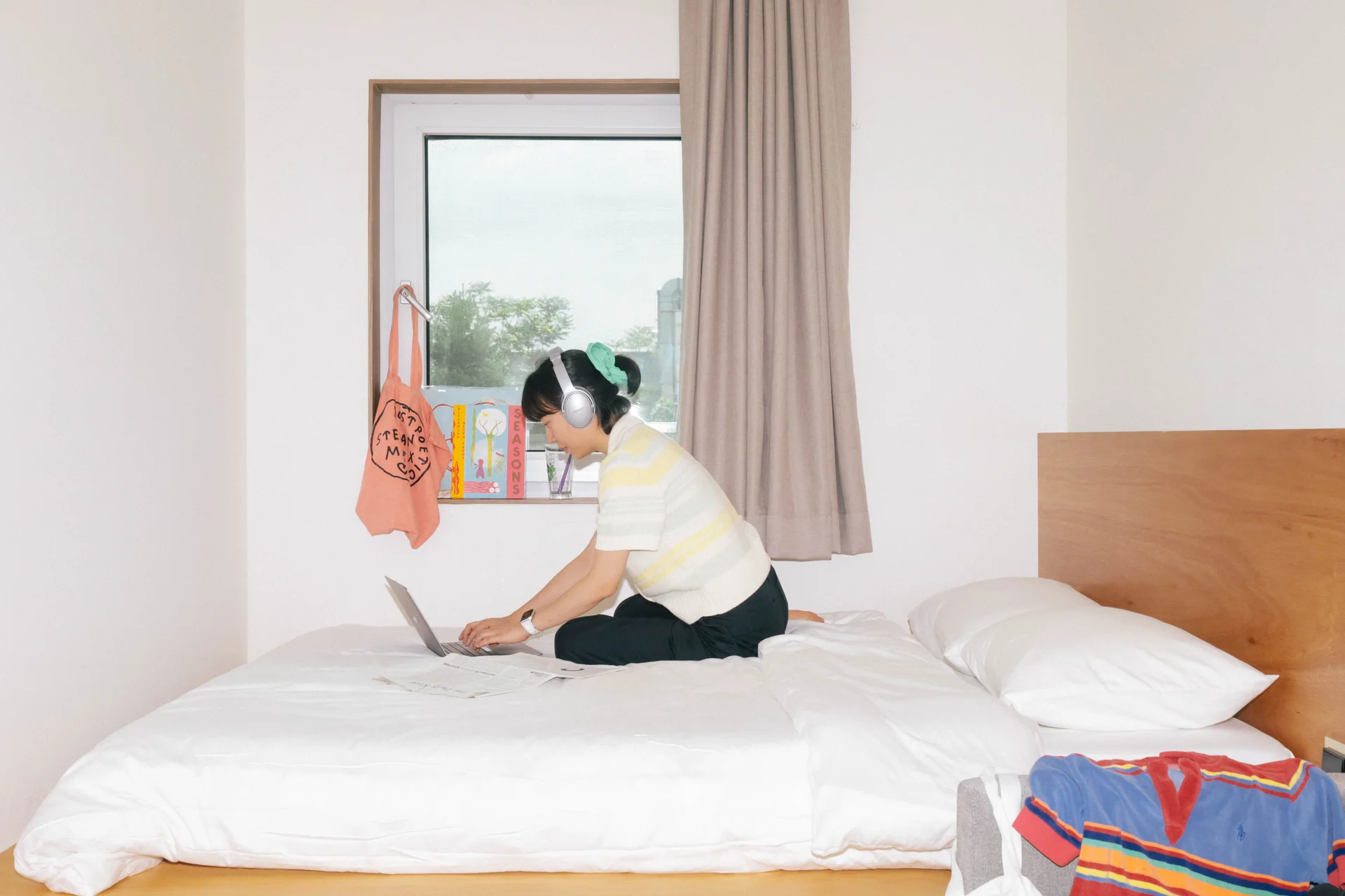 맹그로브 신설 싱글룸에서 한 여성이 침대 위에 앉아 노트북으로 작업하는 모습. 헤드폰을 착용하고 있으며, 창가에는 주황색 가방과 책이 걸려 있고, 침대 위에는 하얀 침구와 옷가지가 놓여 있다.