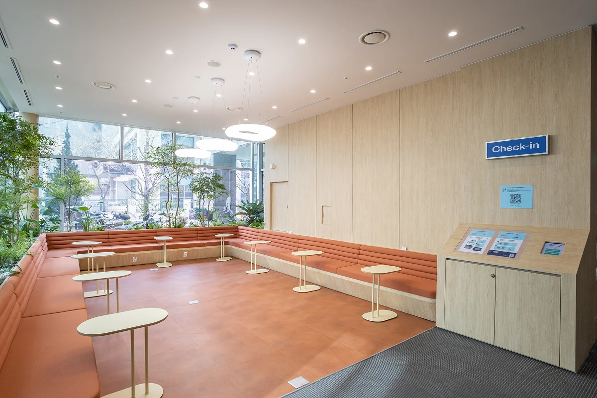 서울 맹그로브 신설 1층 로비 전경. 넓은 통창으로 채광이 가득 들어오며, 따뜻한 톤의 벤치형 좌석과 원형 테이블이 배치된 공간. 입주민과 장기 숙박 이용객이 체크인하거나 대기, 소통할 수 있는 코리빙 하우스 공용 라운지로 쾌적하고 현대적인 분위기를 제공한다.