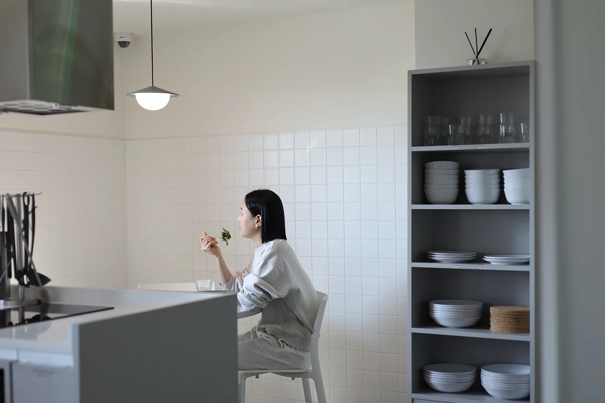 서울 맹그로브 신설 코리빙 하우스 공용 키친. 입주민이 테이블에 앉아 식사를 즐기는 모습으로, 흰색 타일 벽과 정돈된 식기 수납장이 깔끔한 분위기를 연출한다. 장기 숙박과 1인 생활에 적합한 공유 주방으로, 요리와 식사가 가능한 커뮤니티 시설.
