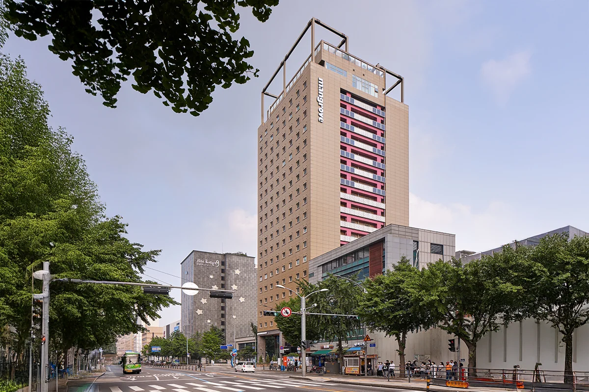 도심 가로수길 옆에 자리한 맹그로브 신설 건물 외관. 베이지 톤의 고층 건물에 커다란 창문과 붉은색 포인트가 돋보인다.