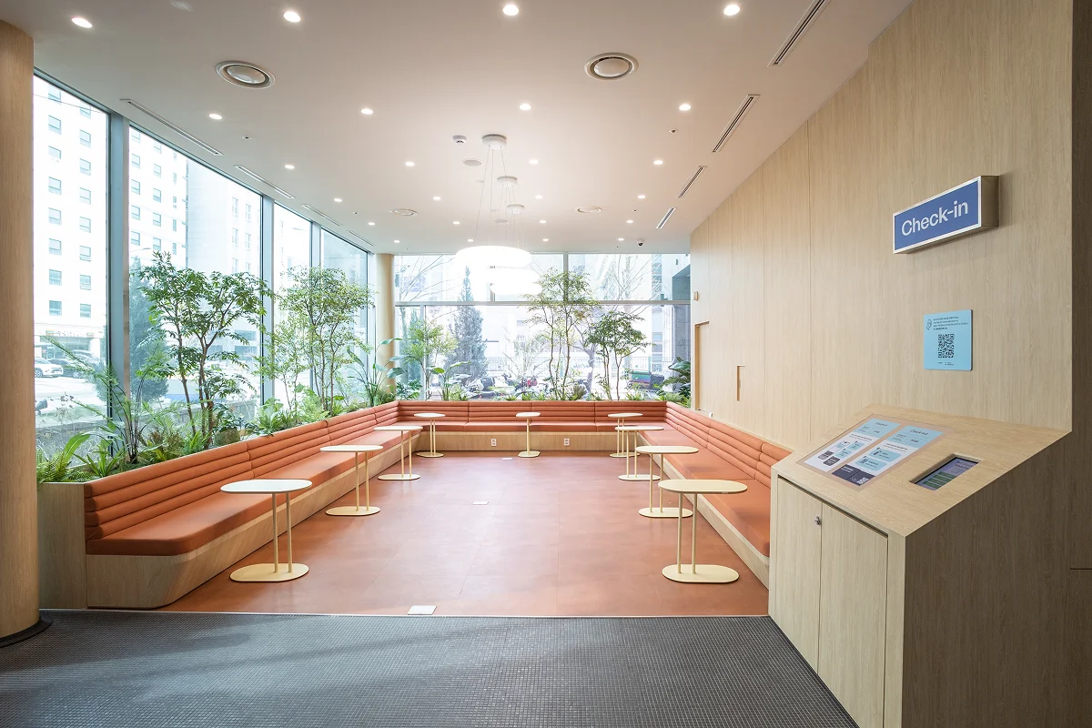 넓은 창으로 자연광이 들어오는 맹그로브 신설 라운지. 오렌지색 소파와 실내 녹색 식물이 조화를 이루며 밝고 쾌적한 분위기를 만든다.