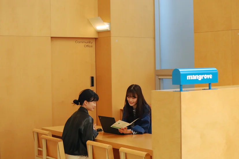 밝은 나무 톤의 인테리어로 꾸며진 맹그로브 동대문 1층 웰컴 라운지. 두 명의 여성이 테이블에 마주 앉아 노트북과 안내 책자를 보며 대화를 나누고 있고, 뒤편에는 ‘Community Office’ 표시가 있으며 오른쪽에는 파란색 맹그로브 로고 간판이 놓여 있다.