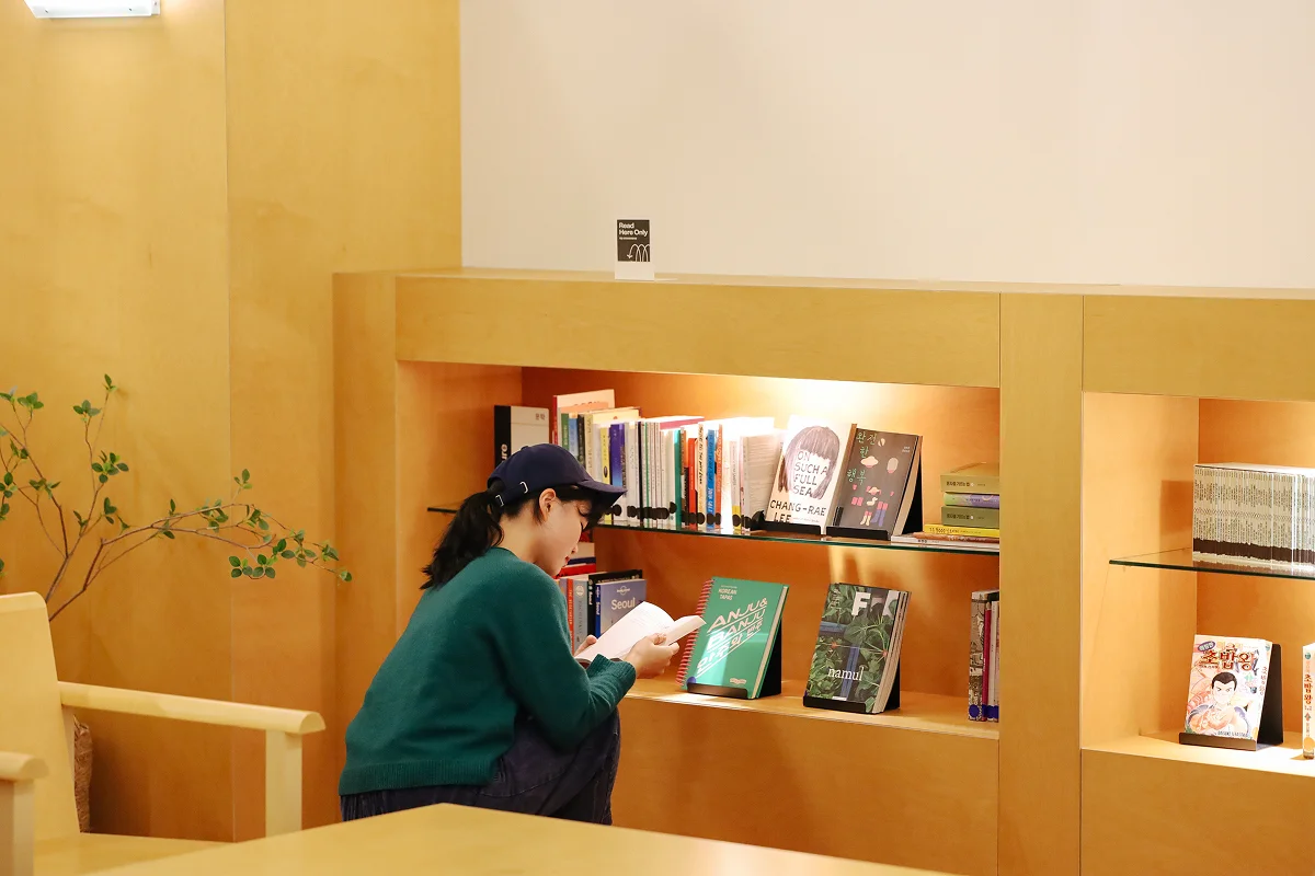 맹그로브 동대문 장기 숙박 B1F 라이브러리 – 따뜻한 우드톤 인테리어의 도서관 공간에서 입주자가 책을 고르는 모습. 장기 체류 고객이 휴식과 독서를 즐기며 머무를 수 있는 아늑한 코리빙 공용 공간.