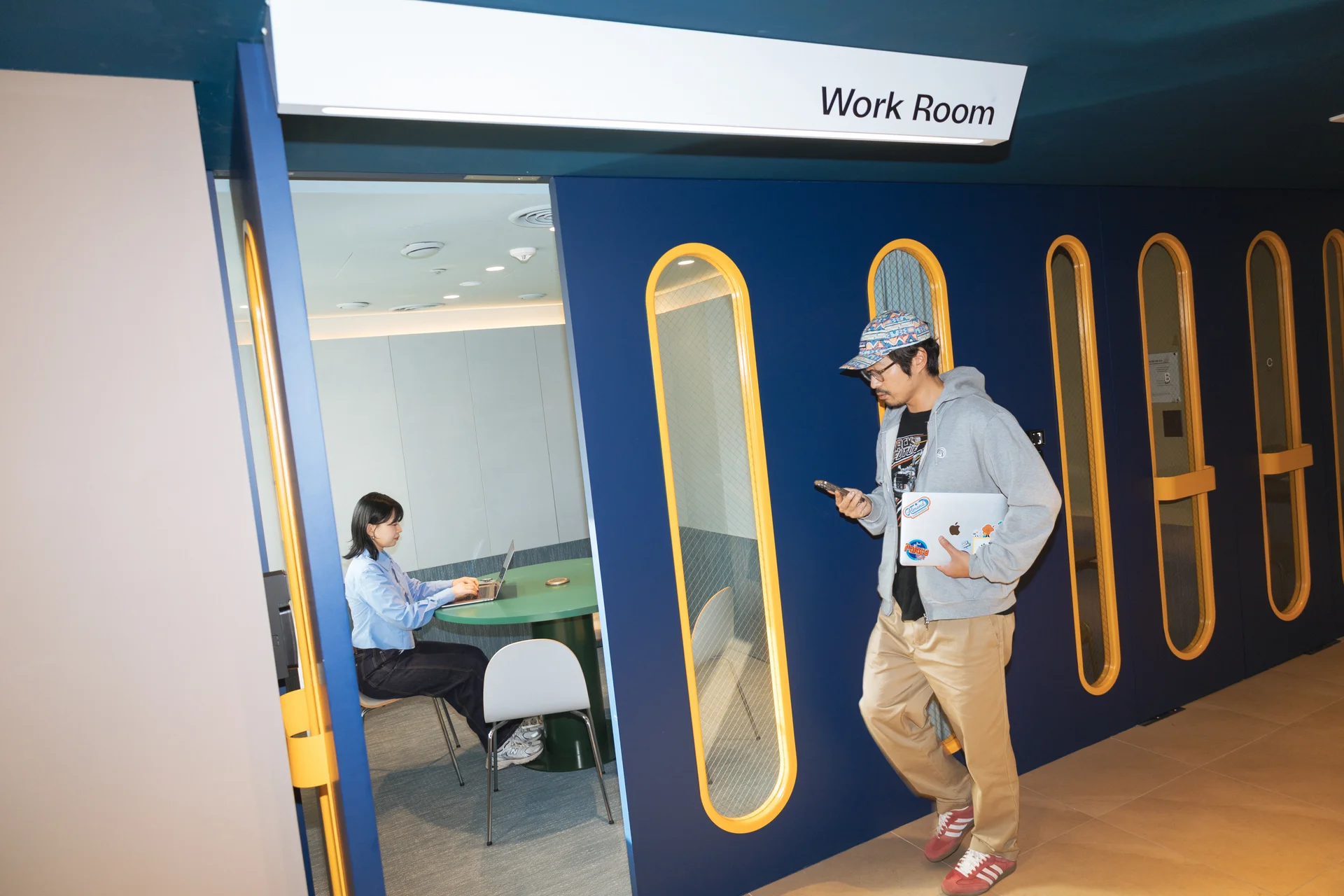 맹그로브 신촌 장기 숙박 5F 워크룸 – 집중할 수 있는 업무 공간에서 입주자가 노트북으로 일하고, 다른 입주자가 자유롭게 이동하는 모습. 장기 체류 고객이 워케이션, 스터디, 회의 등 다양한 용도로 활용할 수 있는 코리빙 전용 공간.