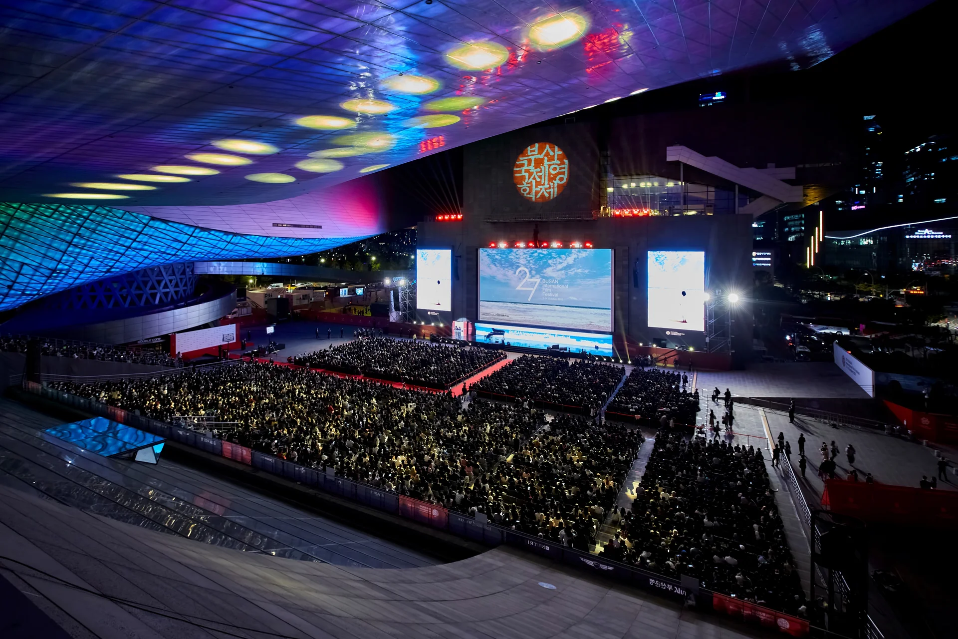 Night view of the Busan International Film Festival (BIFF) opening ceremony at the Busan Cinema Center, with a large seated audience, massive outdoor screen, and vibrant lighting under the futuristic roof structure.