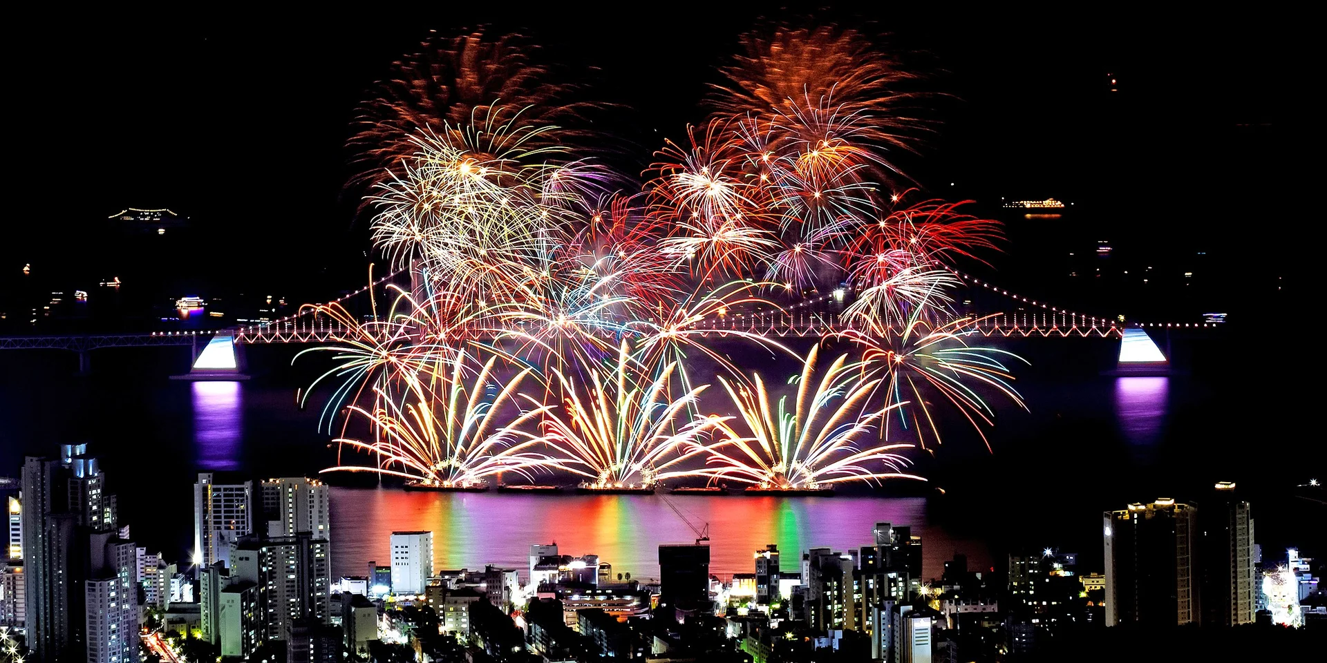 Spectacular fireworks display over Gwangalli Beach and Gwangan Bridge during the Busan International Fireworks Festival, with vibrant colors reflected on the sea and city skyline illuminated at night.